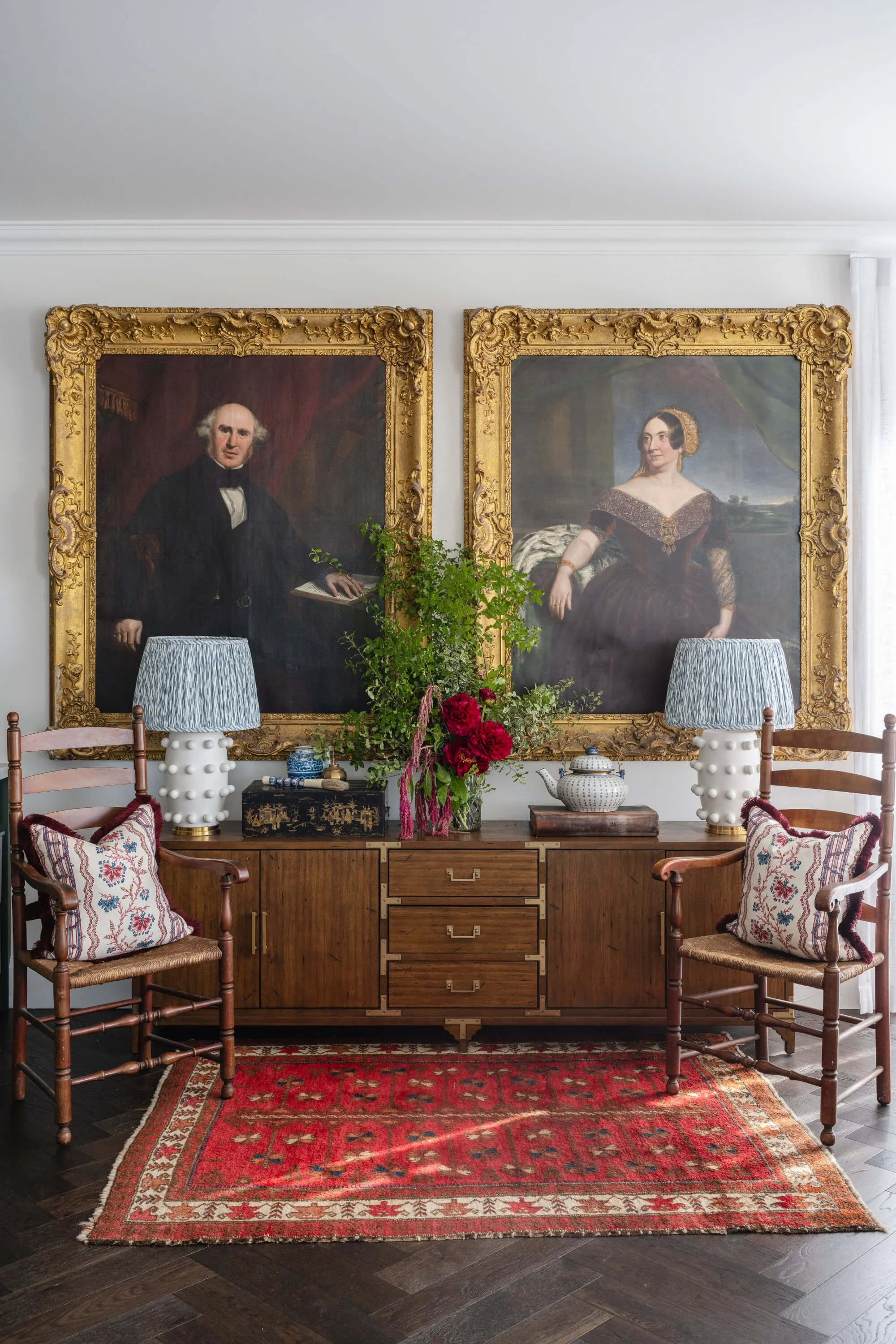 Interior of a room with two large framed portraits of historical figures hanging on a white wall. There is a wooden sideboard with a floral arrangement, a teapot, and small decorative objects. Ascot Residential Interior Design by Biju Interiors.
