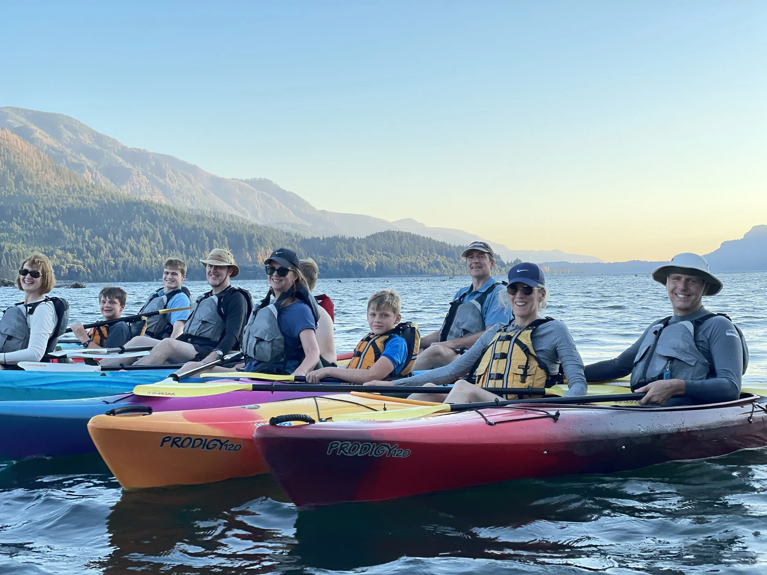 A group of people sitting in colorful kayaks on a lake, smiling with mountains and a clear sky in the background.