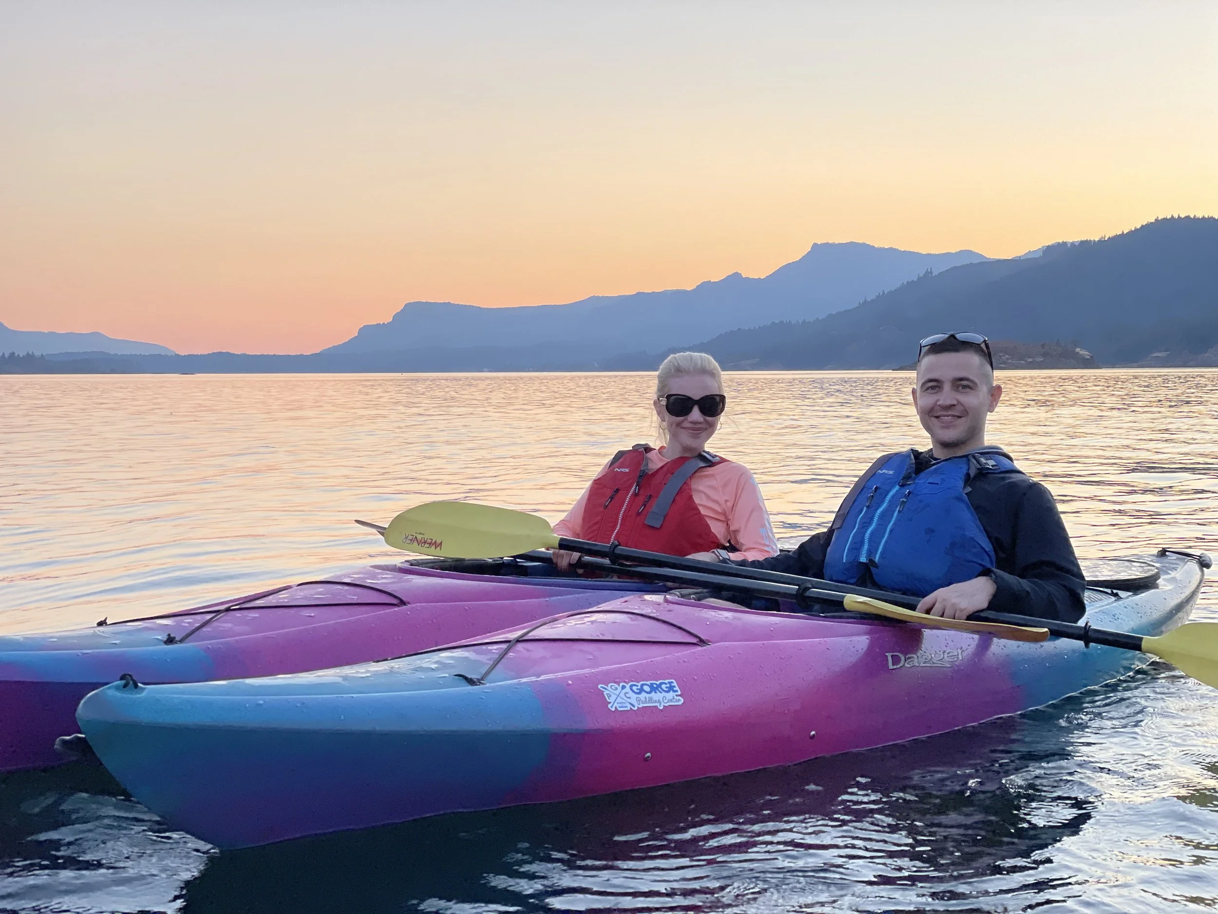 Two people kayaking in water at sunset, with mountains and a colorful sky in the background.