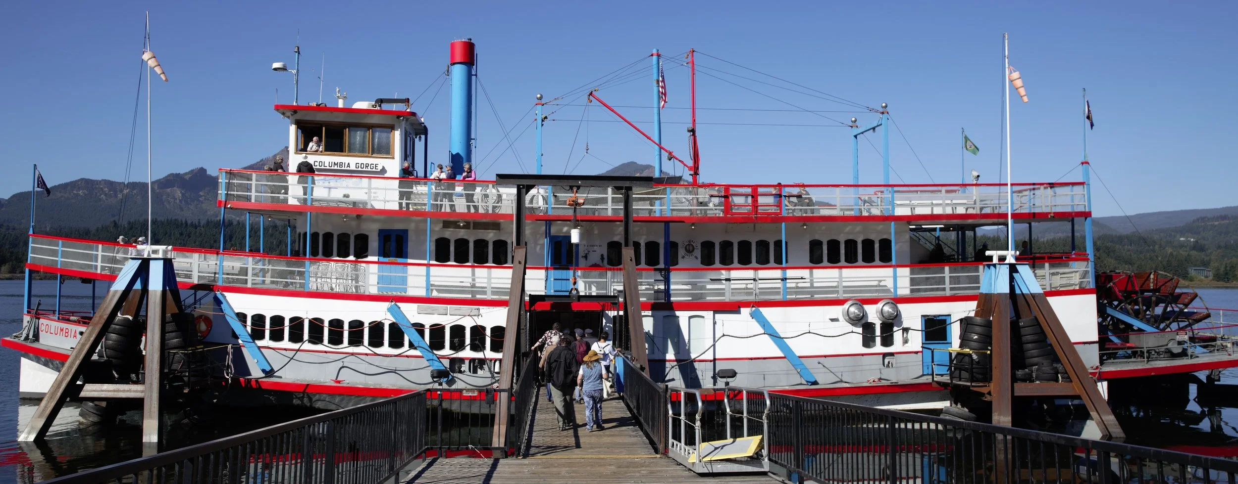An old-fashioned paddleboat named Columbia Gorge docked at a pier with people boarding, mountains in the background, under a clear blue sky.