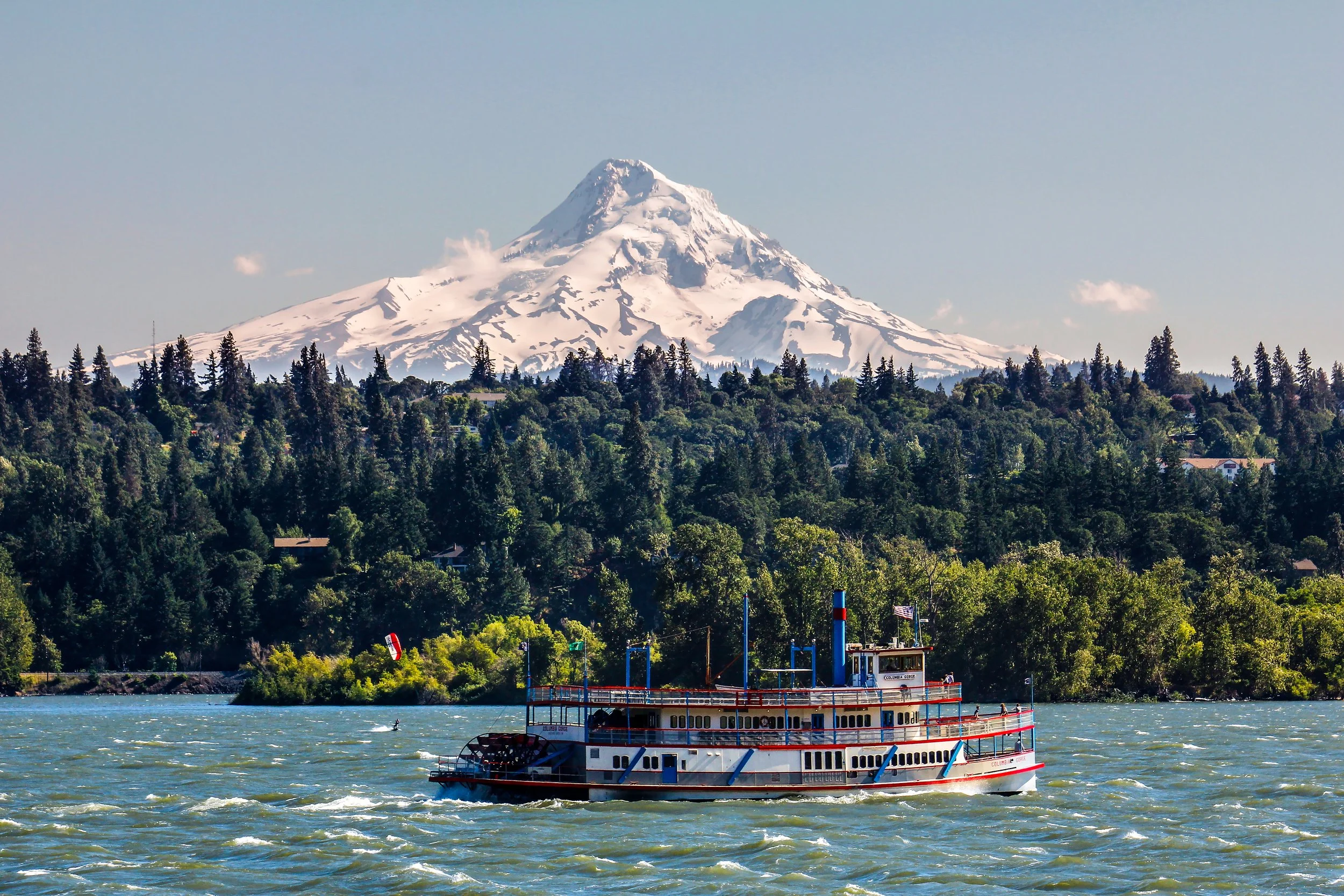 A boat on a body of water near a forested shoreline with a snow-capped mountain in the background.