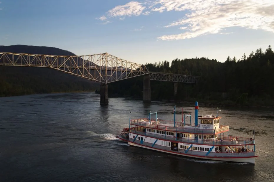 A ferry boat painted in white with red and blue accents traveling under a large metal bridge over a river with forested hills in the background during sunset.