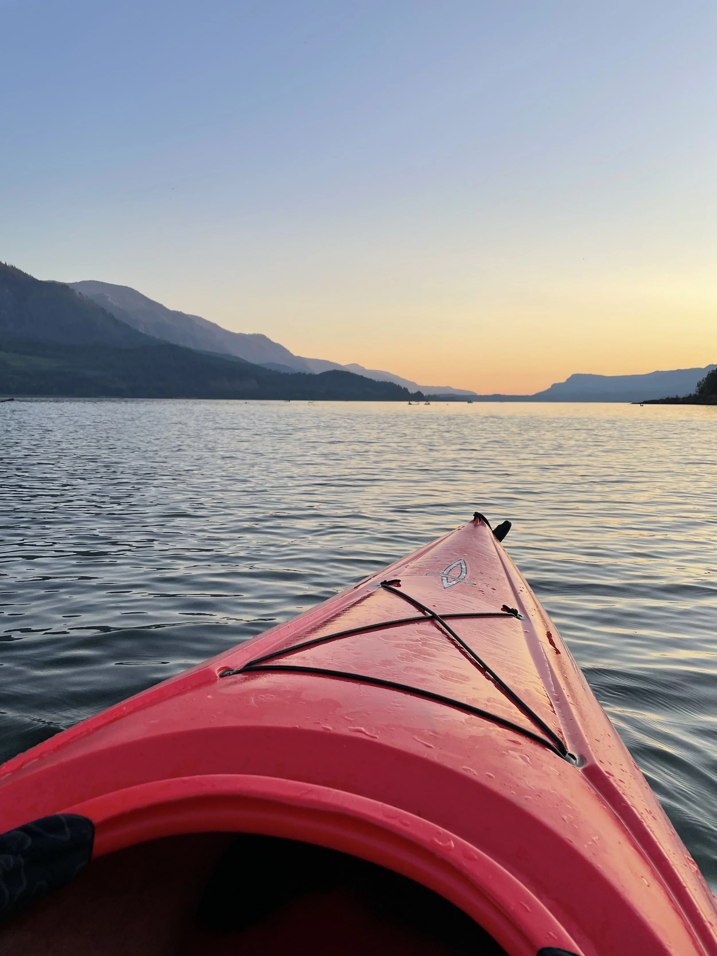 Looking from the front of a pink kayak at a river or lake during sunset, with mountains in the background and small boats on the water.