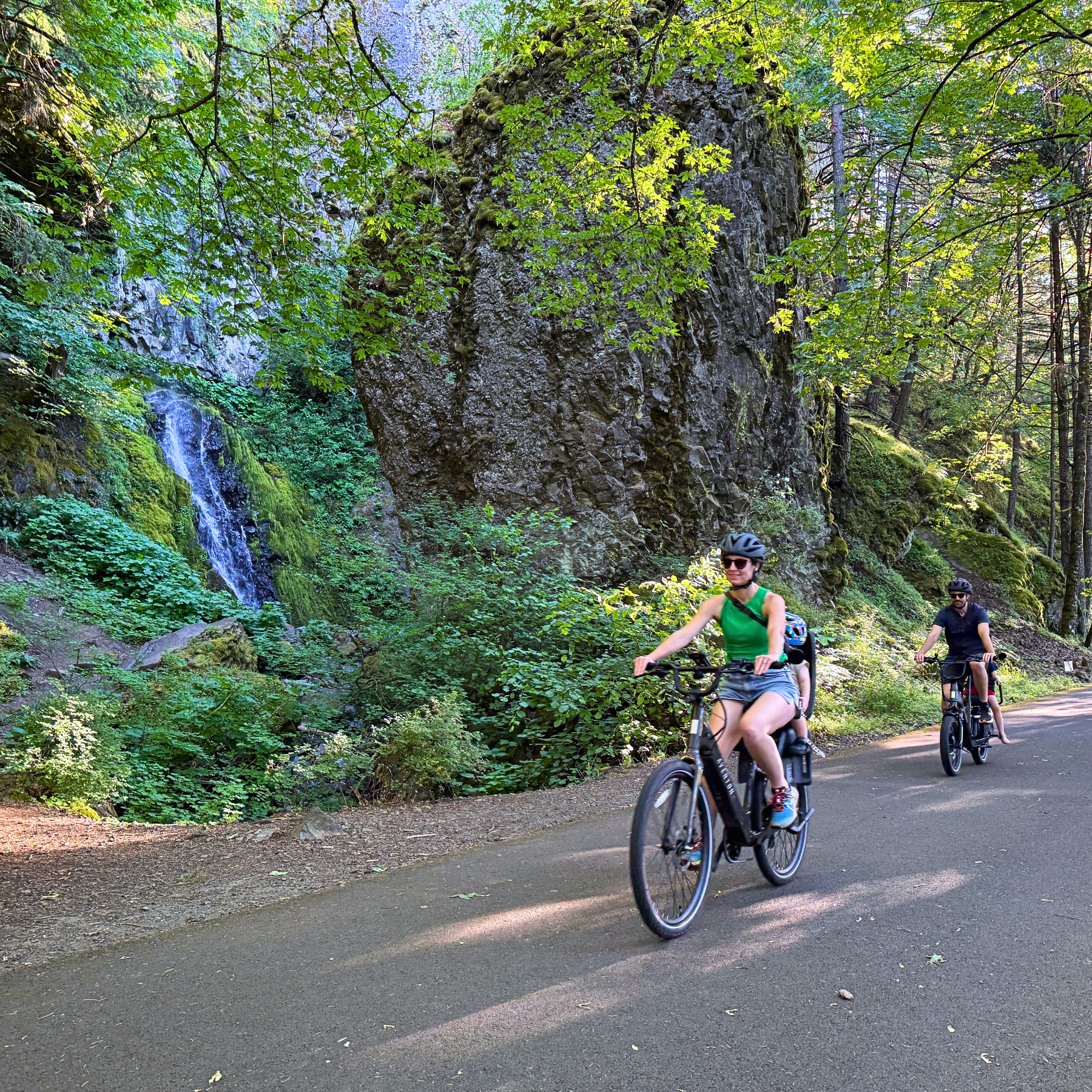 Two women riding bicycles down a forest trail surrounded by green trees, moss-covered rocks, and a small waterfall.