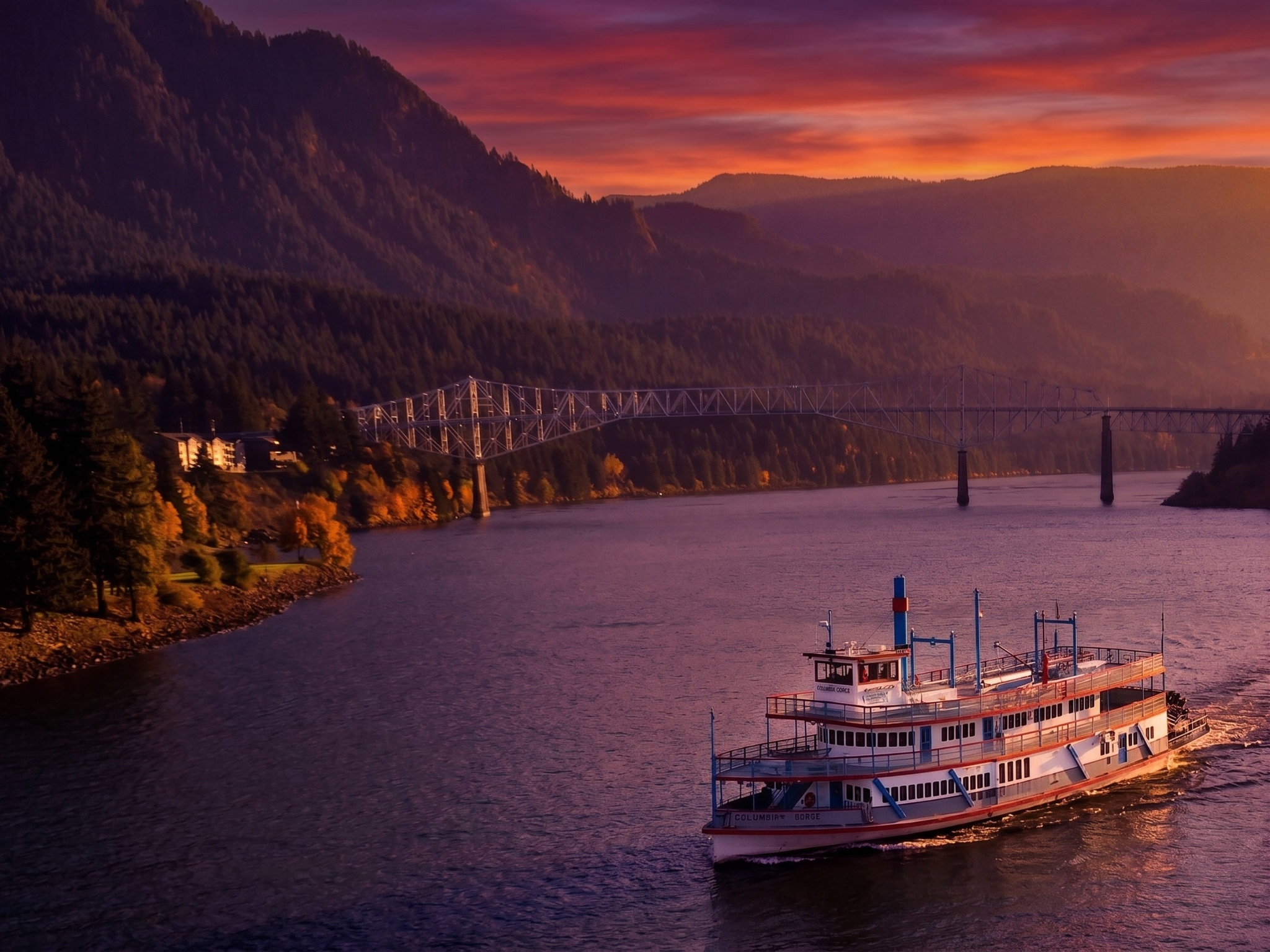 A scenic view of a river at sunset with mountains in the background, a bridge spanning across the river, and a ferry boat in the foreground.