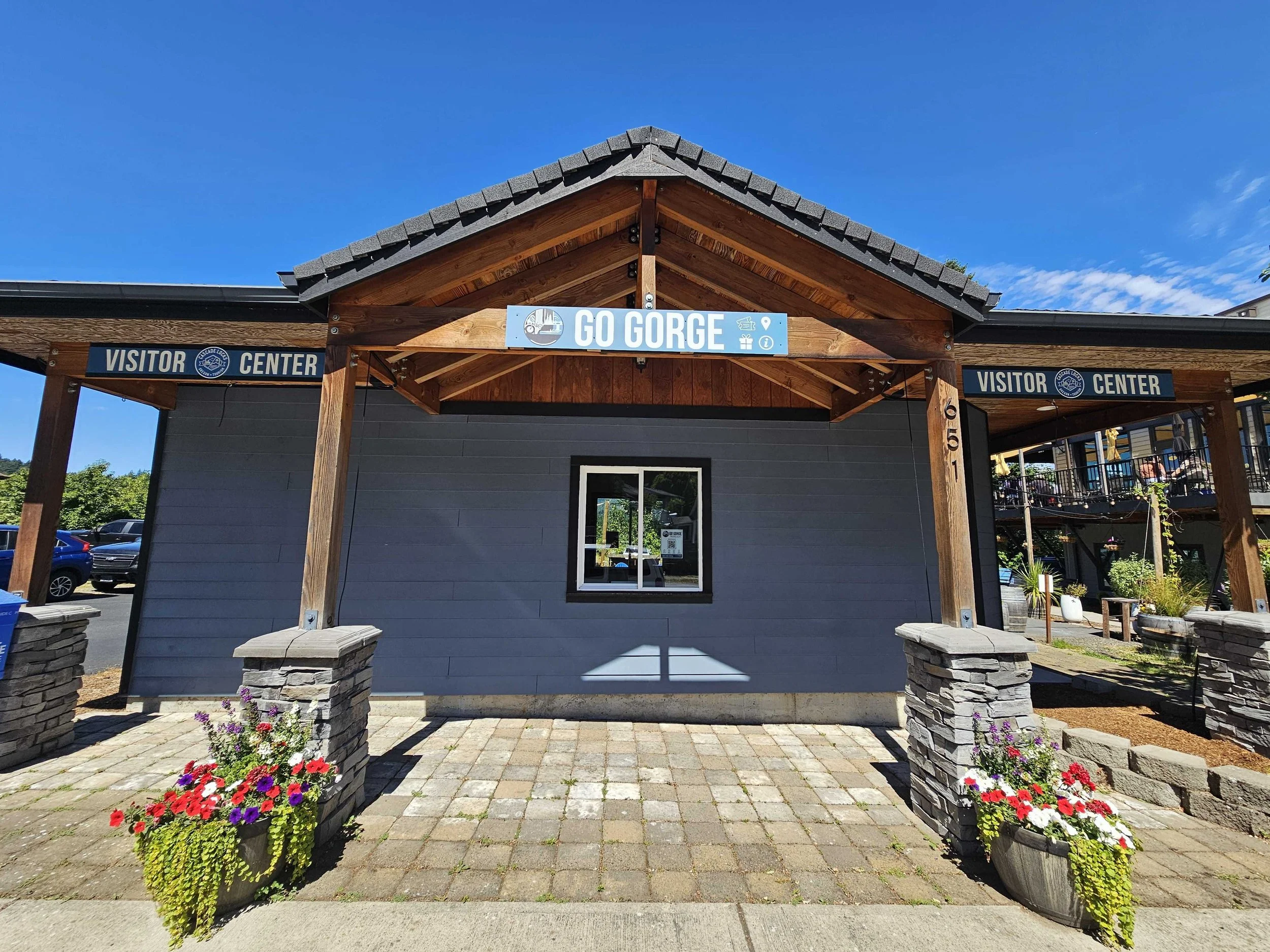 Wooden structure with sign that says 'Go Gorge' at the entrance of a visitor center, with two flower planters at the front and a parking lot with cars visible in the background.