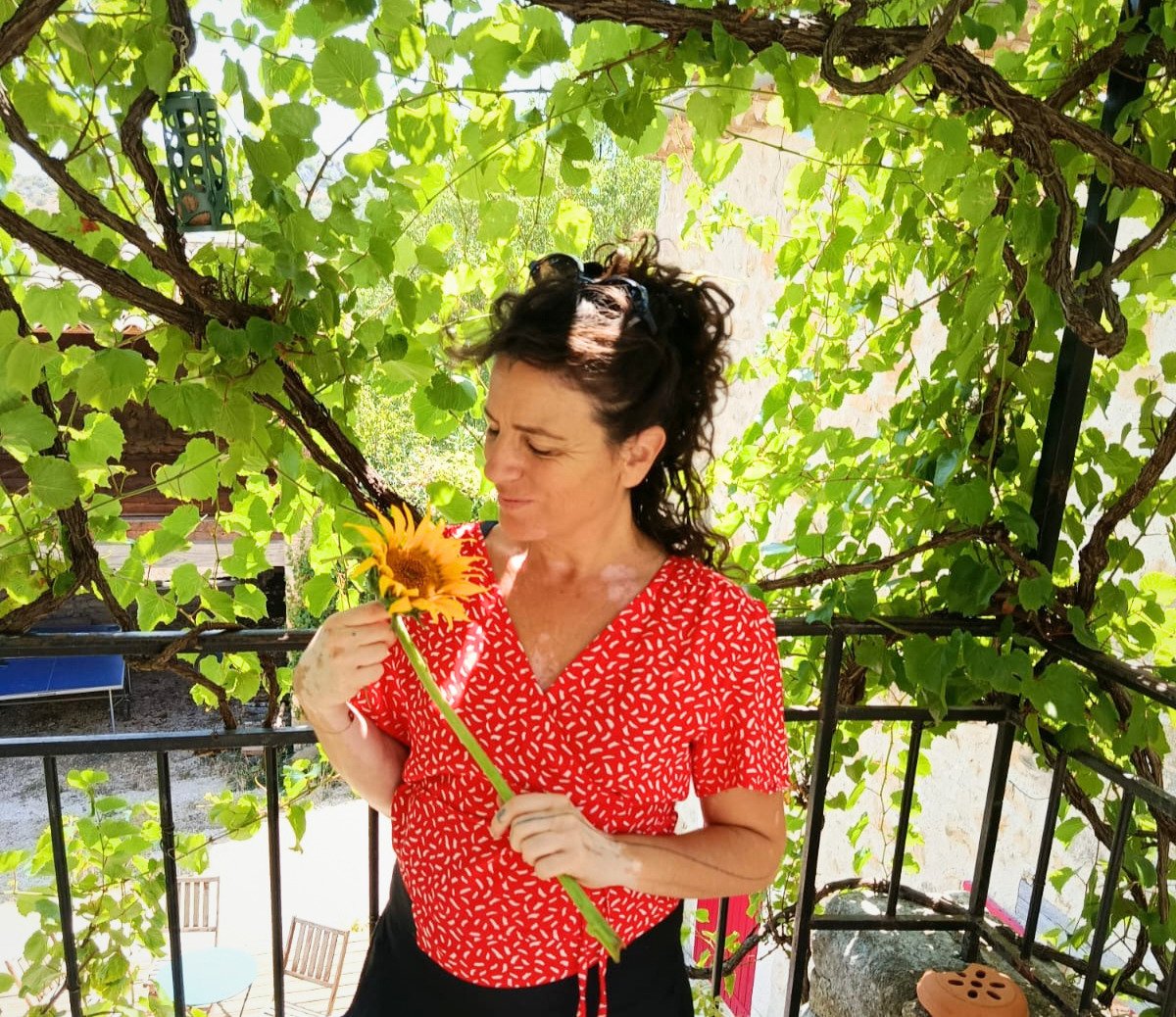 Woman holding a sunflower, standing on a balcony surrounded by green vines and leaves.