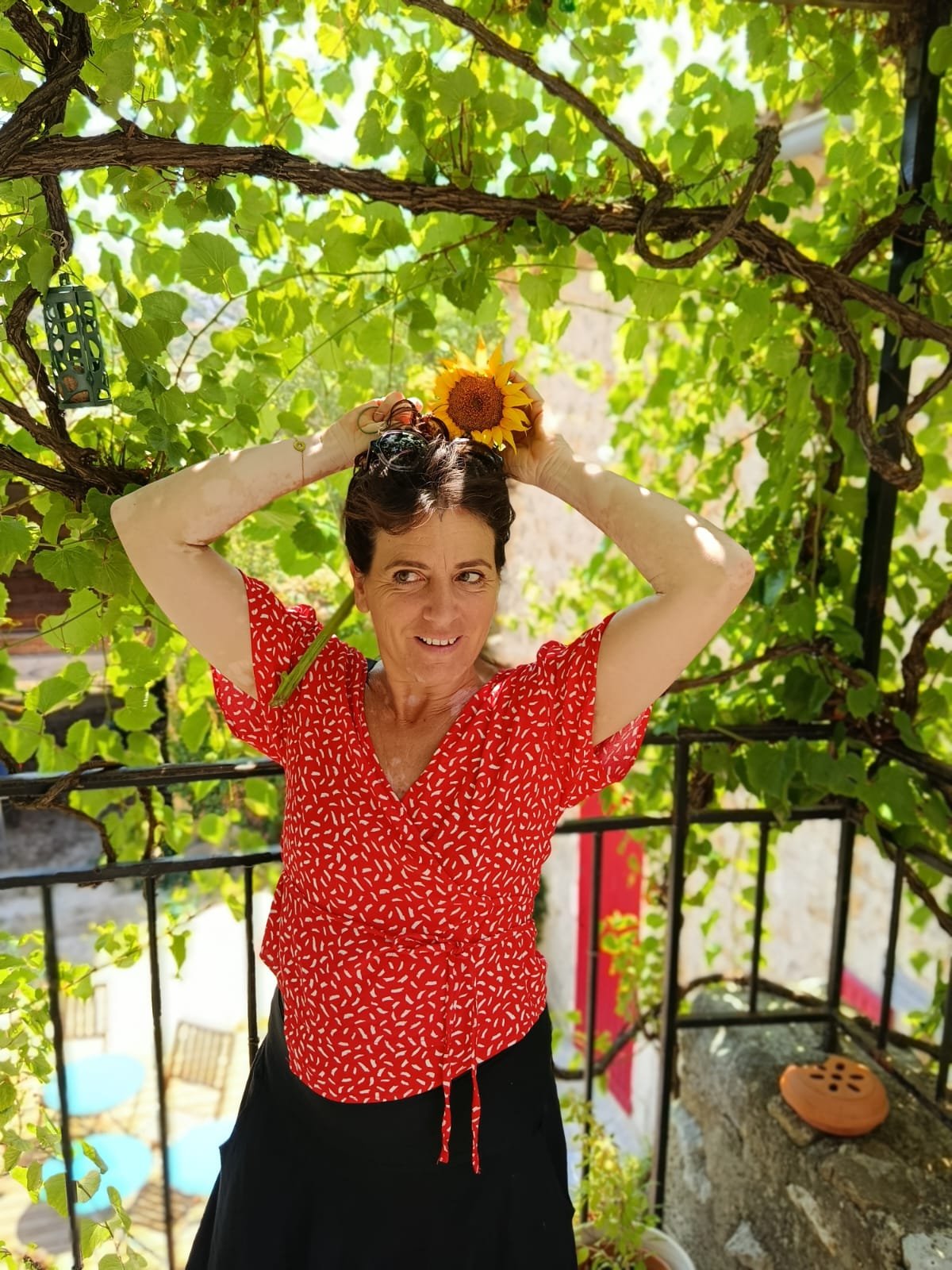 Femme souriante portant une robe rouge avec un motif blanc, tenant une fleur de tournesol sur la tête, dans un jardin ombragé avec des feuilles vertes et un hélicoptère en plastique suspendu à la branche d'un arbre.