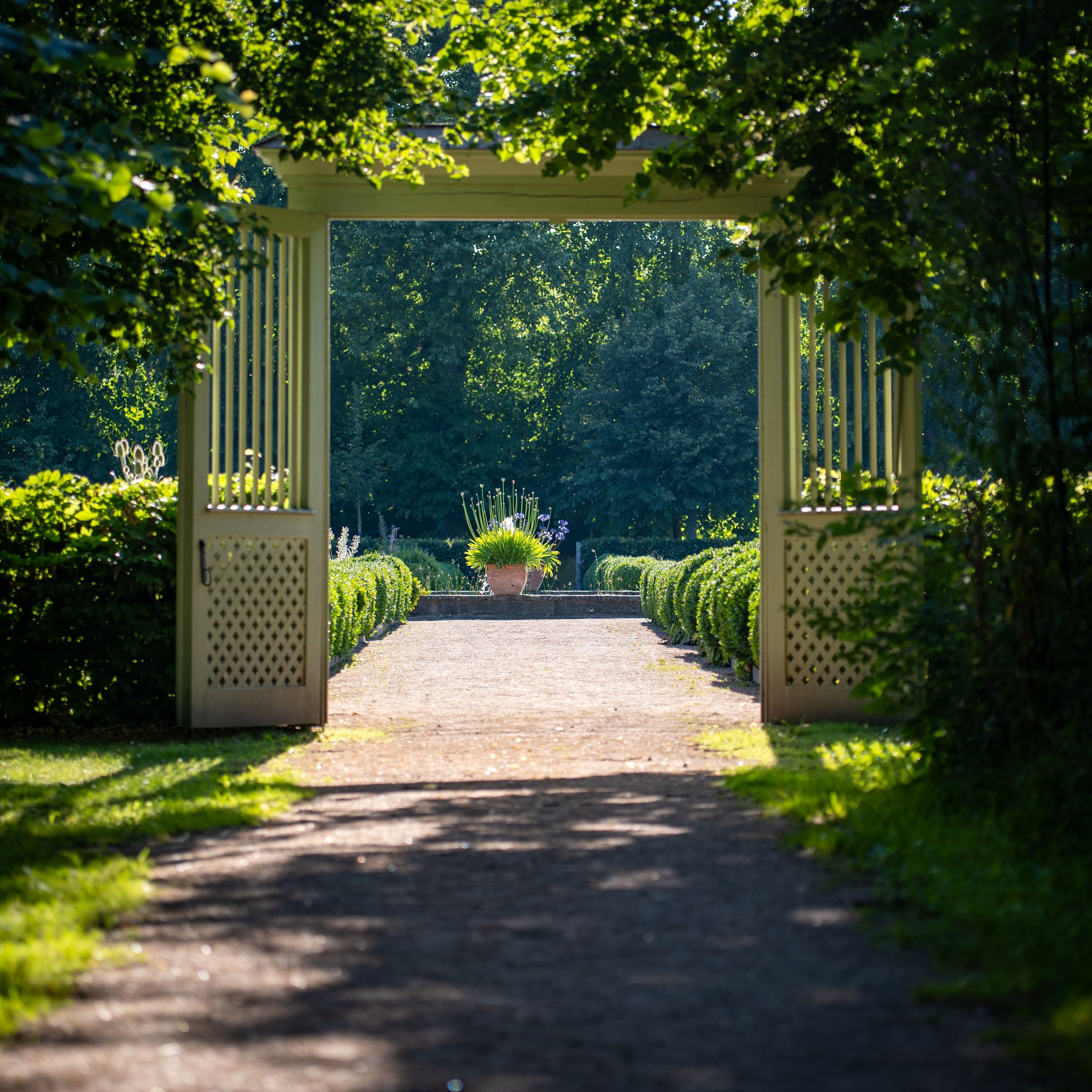 Un portail en bois blanc ouvert, entouré de végétation verte luxuriante, avec des arbres et un jardin fleuri visible au fond, sous la lumière du soleil.