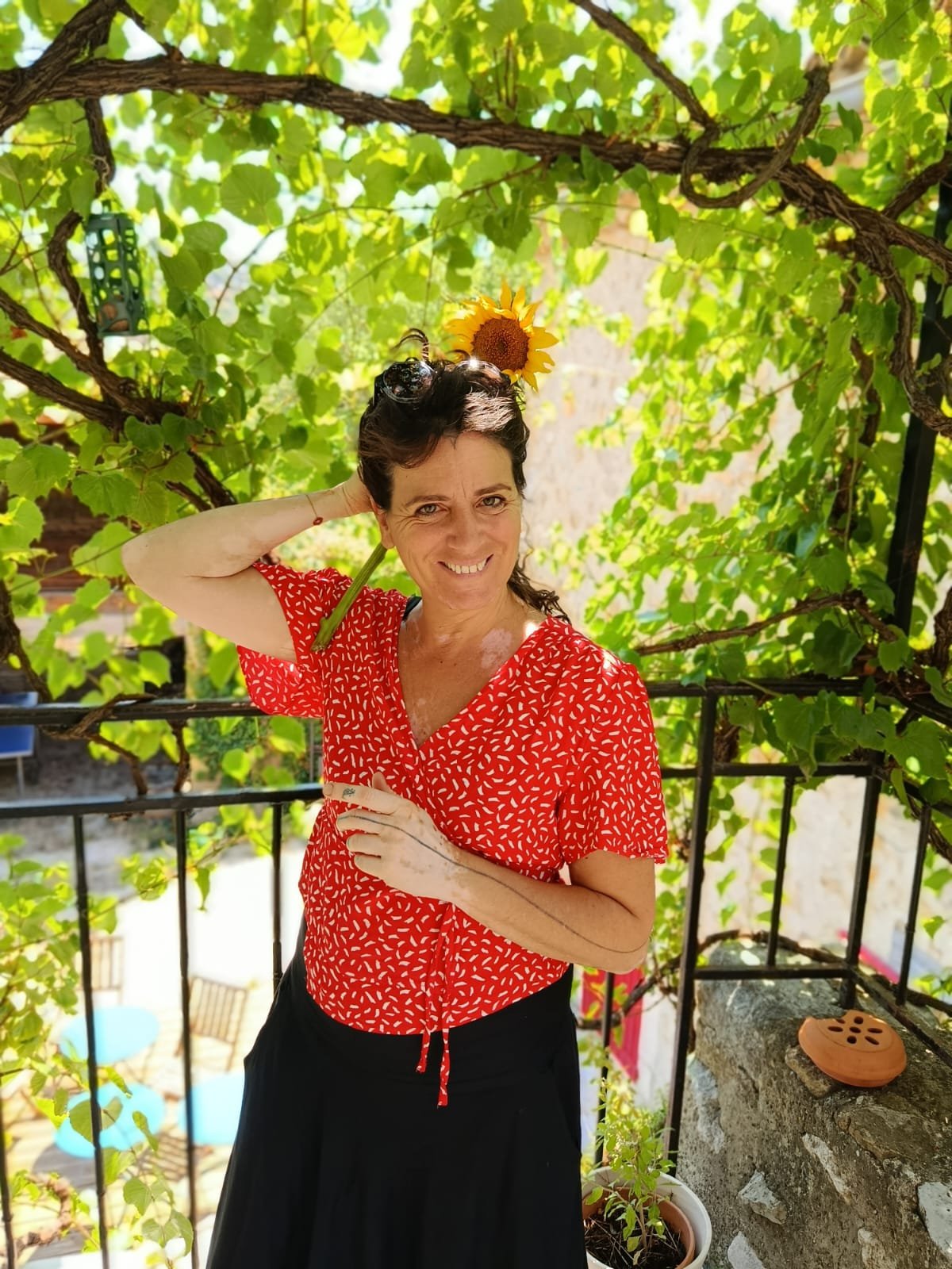 Une femme souriante portant une robe rouge à motifs blancs, tenant une branche de vigne sur la tête avec une fleur de tournesol, se tenant devant un treillis de vigne vert.