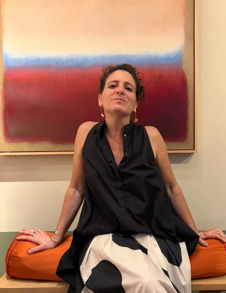 Femme assise sur un banc avec un coussin orange, portant une robe noire, devant une peinture abstraite aux couleurs chaudes.