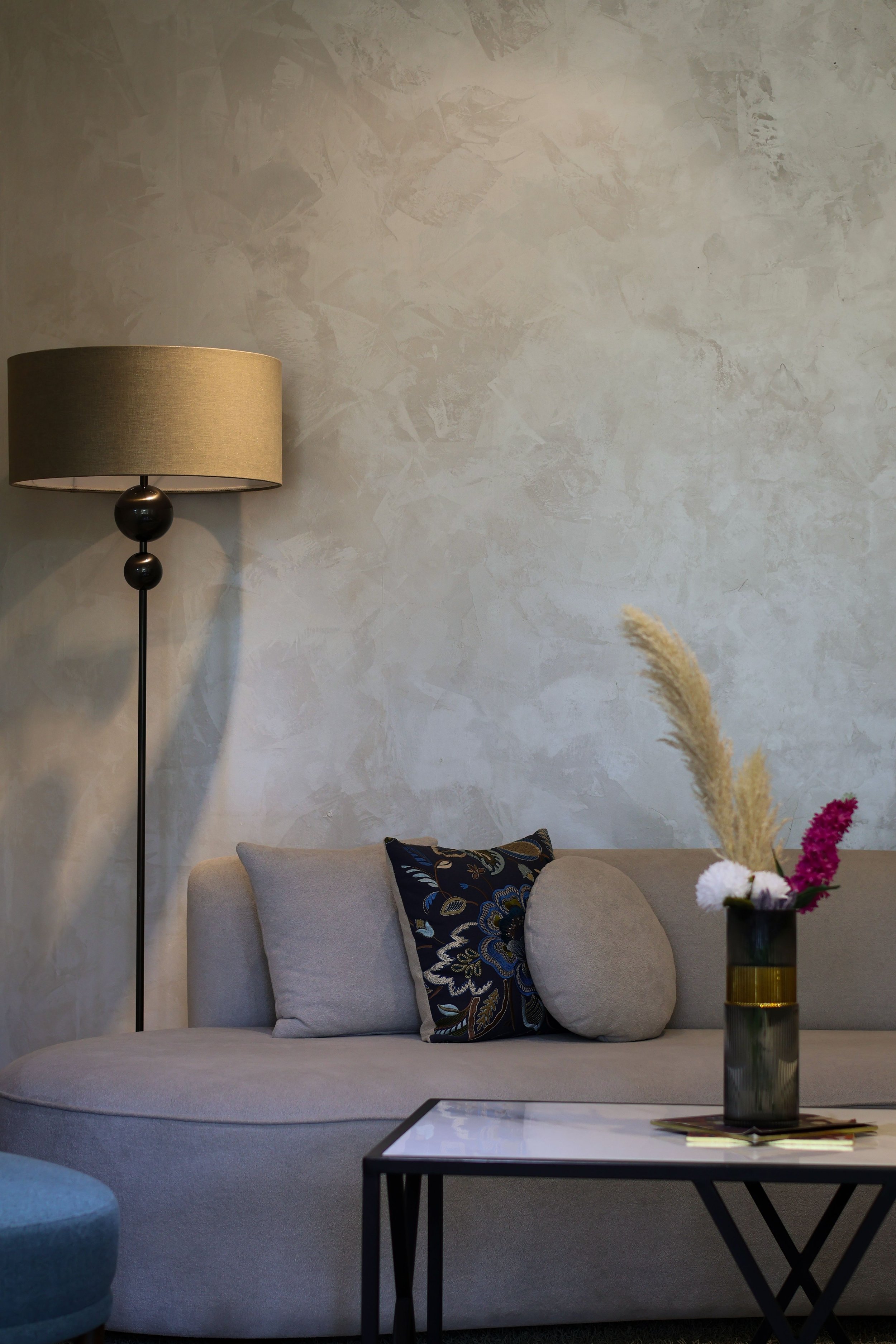 Intérieur d'un salon moderne avec un canapé beige, coussin décoratif avec un motif floral, lampadaire au style contemporain, table basse avec vase contenant des fleurs, mural en plâtre et tapis de sol.