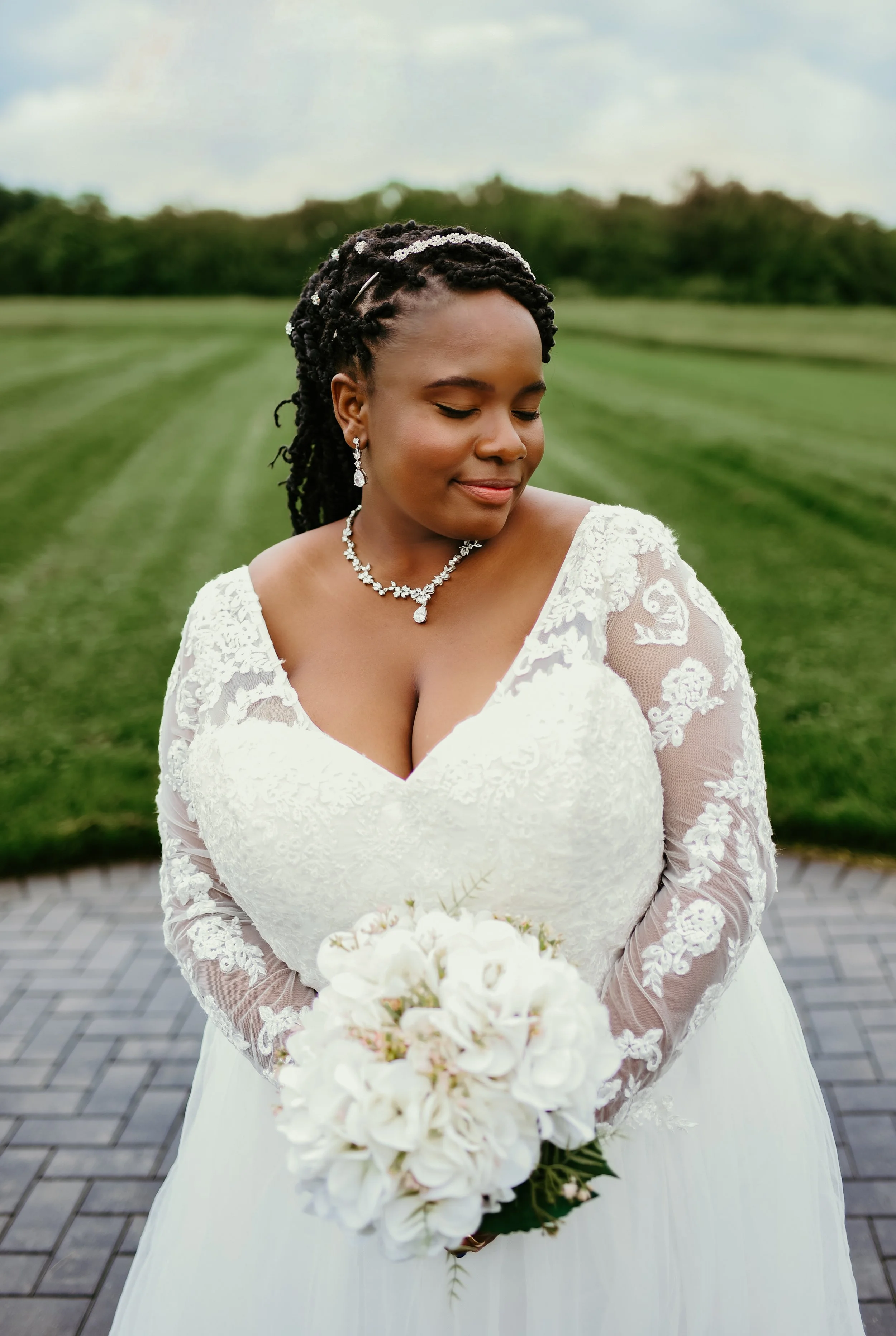 A bride in a white lace wedding dress with long sleeves, holding a bouquet of white flowers, and standing outdoors on a paved walkway with green grass and trees in the background, with her eyes closed and a gentle smile.