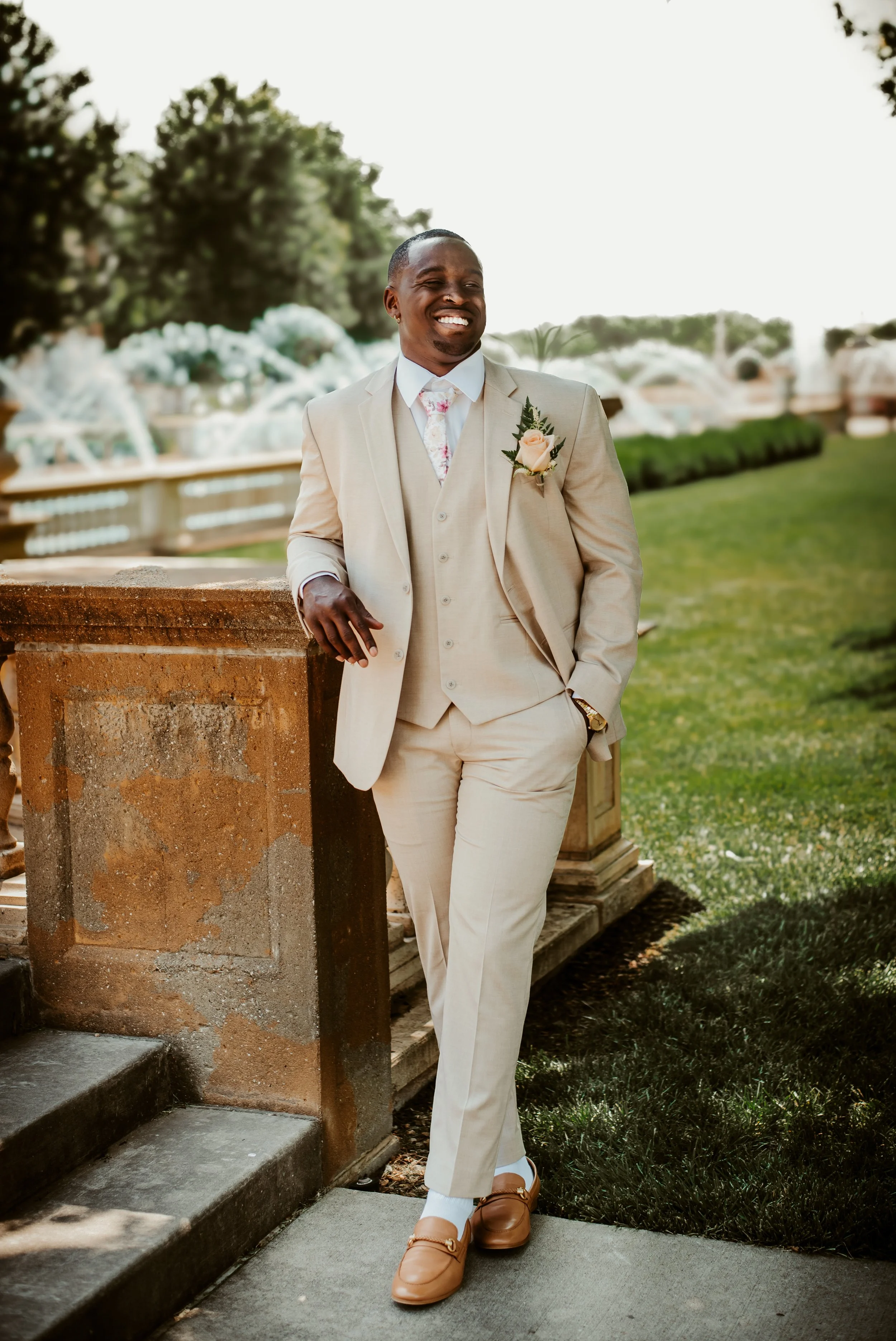 A man in a light beige suit, white shirt, patterned tie, and tan dress shoes standing outdoors near a brick ledge and fountain, smiling and looking to his right.