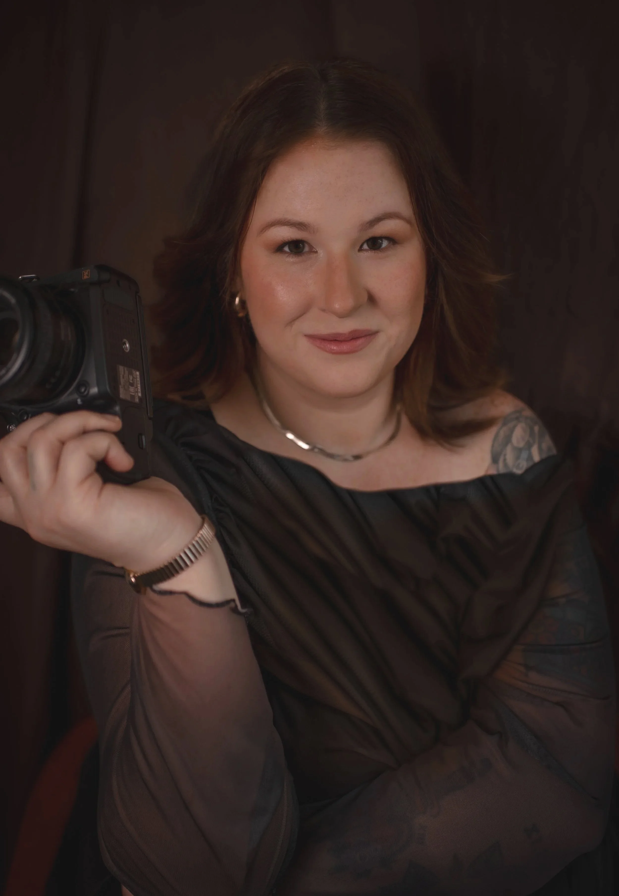 A woman with shoulder-length brown hair holding a camera, wearing a dark top, sitting on the floor with a dark background.