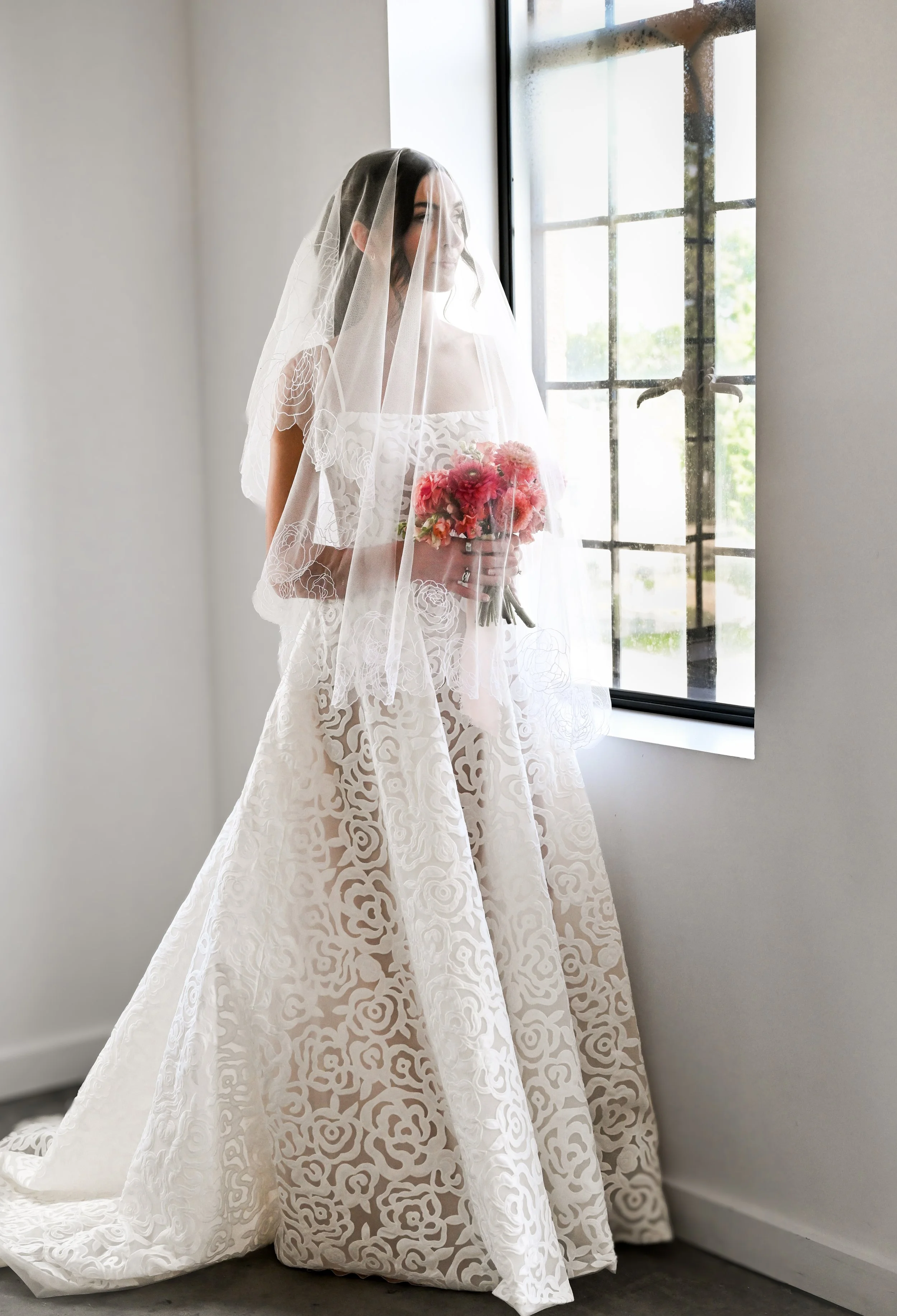 A bride in a white lace wedding dress with floral patterns, holding a bouquet, standing by a window with sunlight coming through.