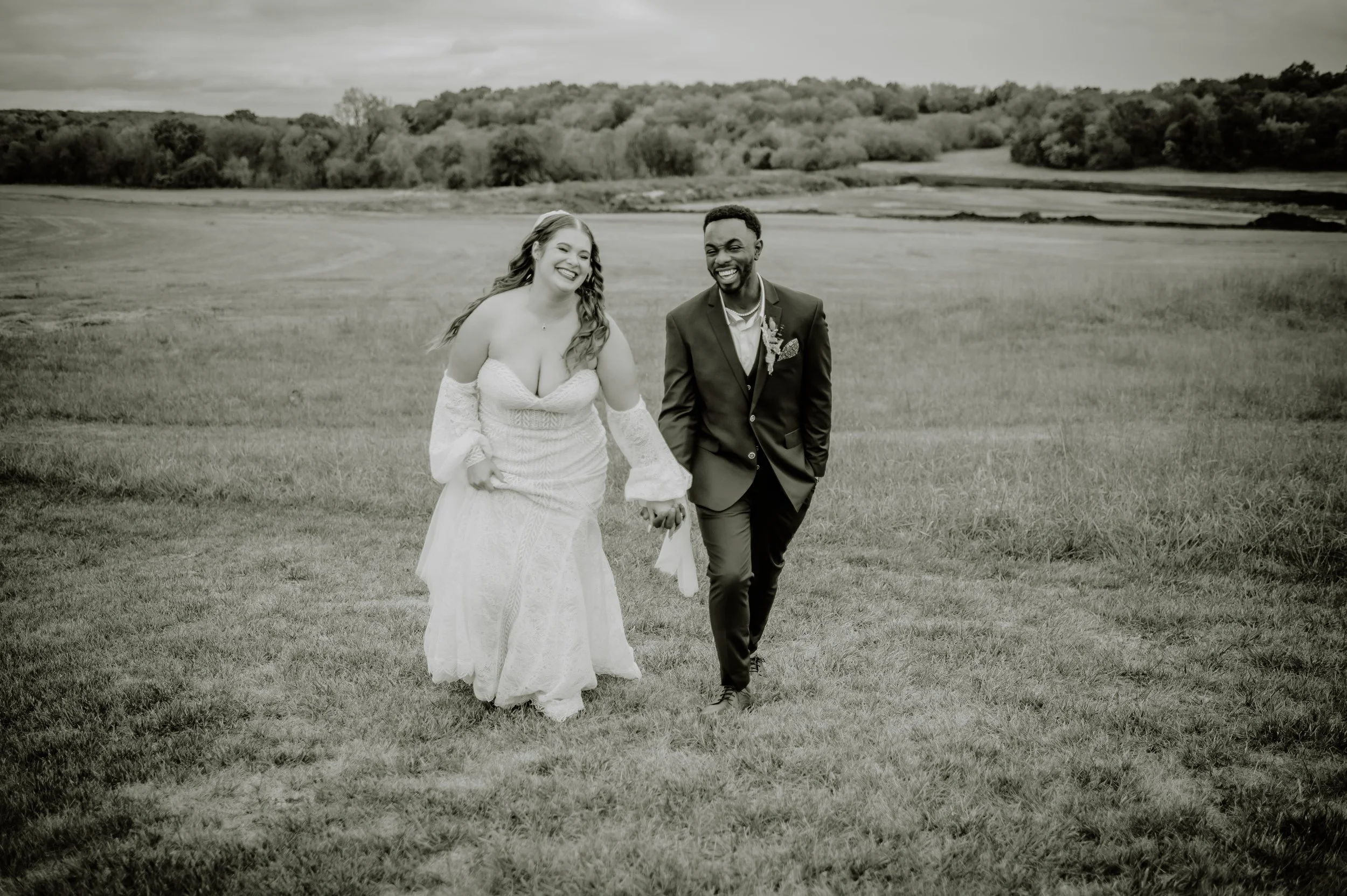 A smiling bride and groom walking hand in hand outdoors on a grassy field with hills and trees in the background, in black and white.