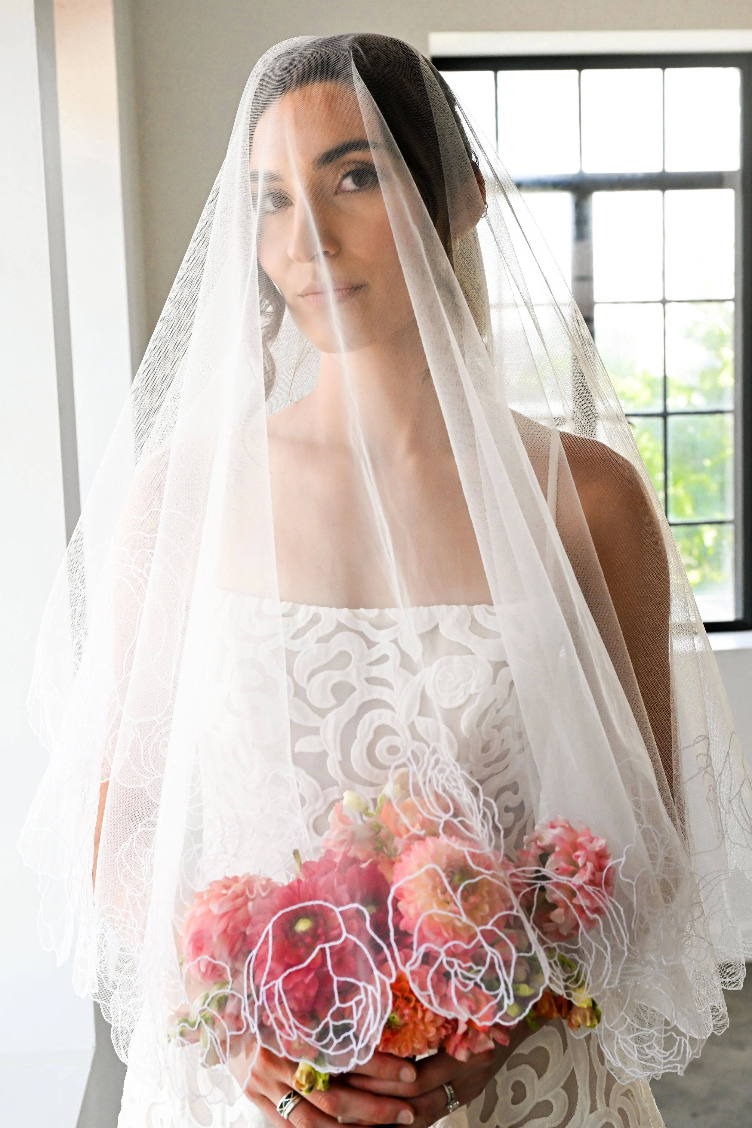 A woman dressed as a bride, wearing a white wedding dress and veil, holding a bouquet of pink and red flowers, standing in front of a window.