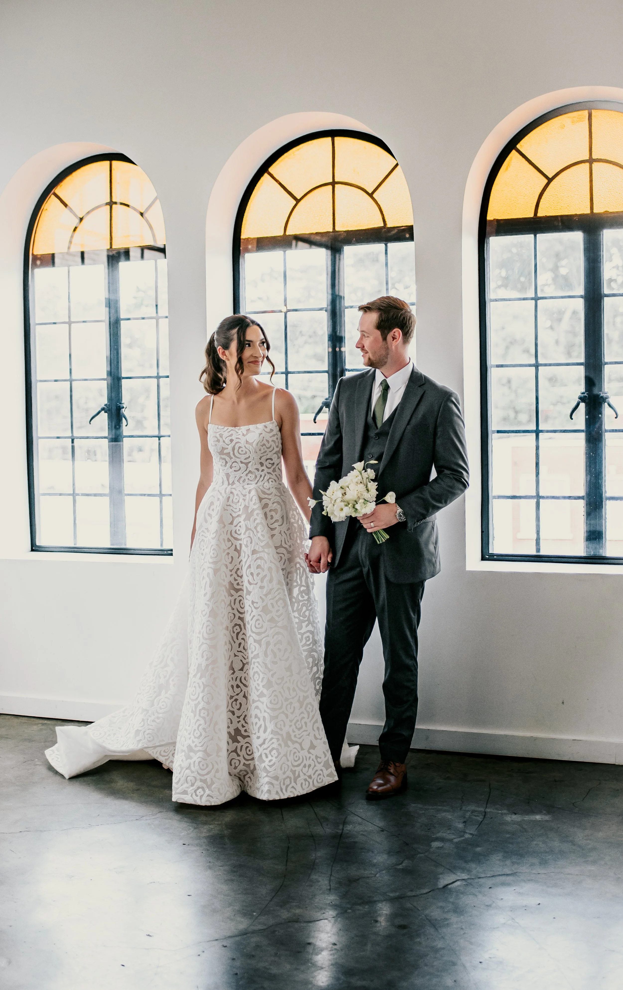 A bride and groom standing hand in hand in front of large arched windows, with soft natural light illuminating their faces. The bride is wearing a white wedding gown with intricate swirling patterns, and the groom is in a dark suit and green tie, hol