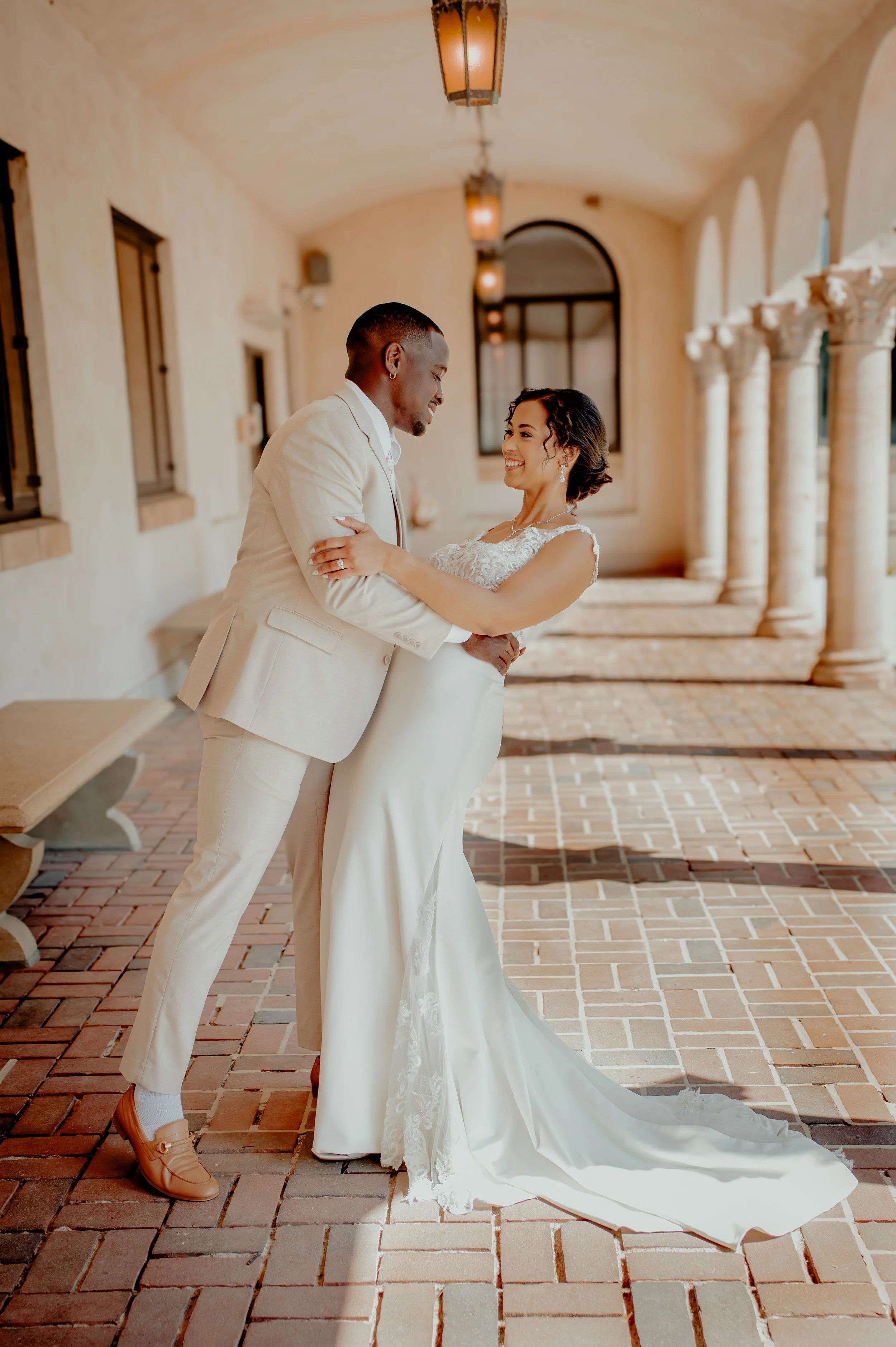 A bride and groom sharing a dance or moment together outdoors on a brick walkway, with the bride in a wedding dress and the groom in a light-colored suit.