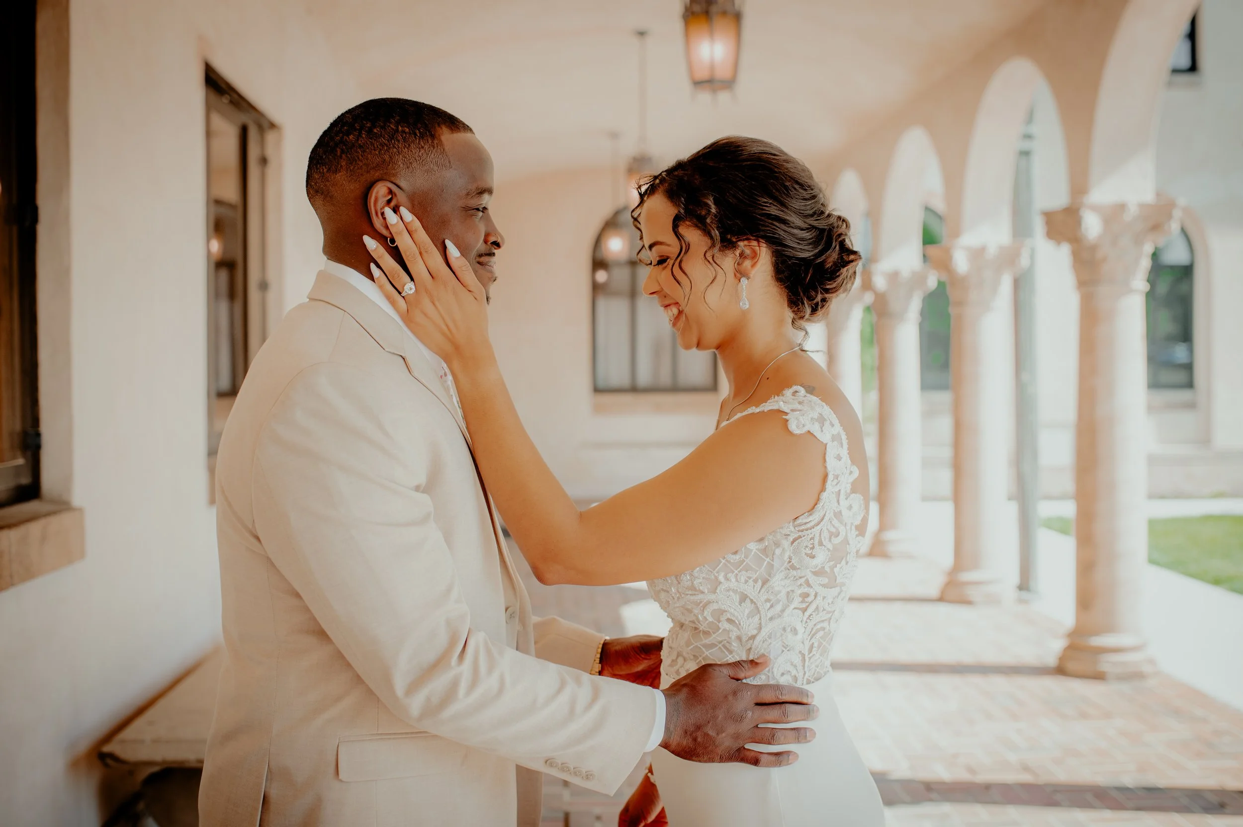 A bride and groom sharing a tender moment indoors, with the bride smiling and touching the groom's face, both wearing wedding attire, in a venue with arches and large windows.
