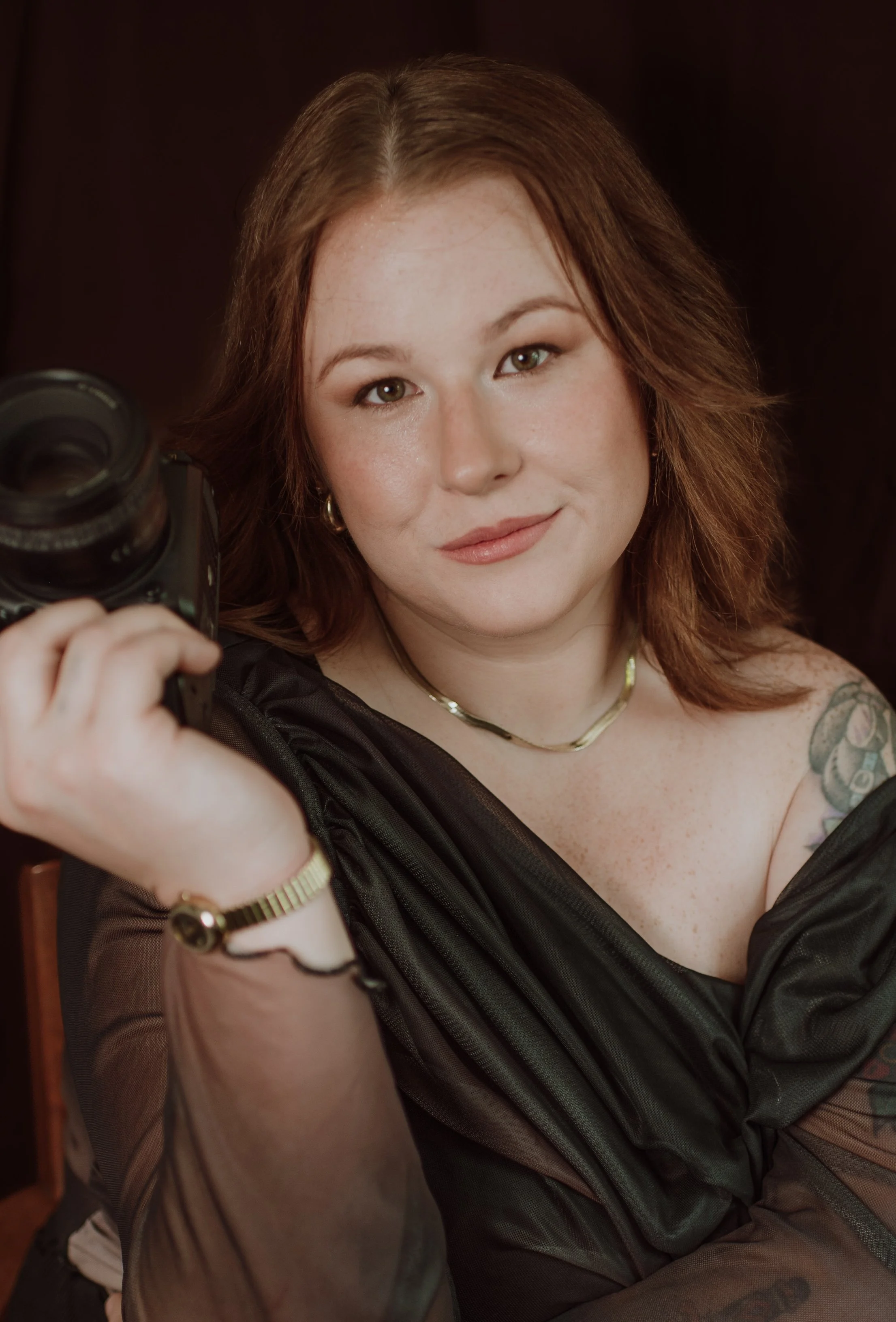A woman with red hair and light skin takes a selfie, holding a camera with a focused expression, wearing a black outfit, jewelry, and has tattoos on her arm.