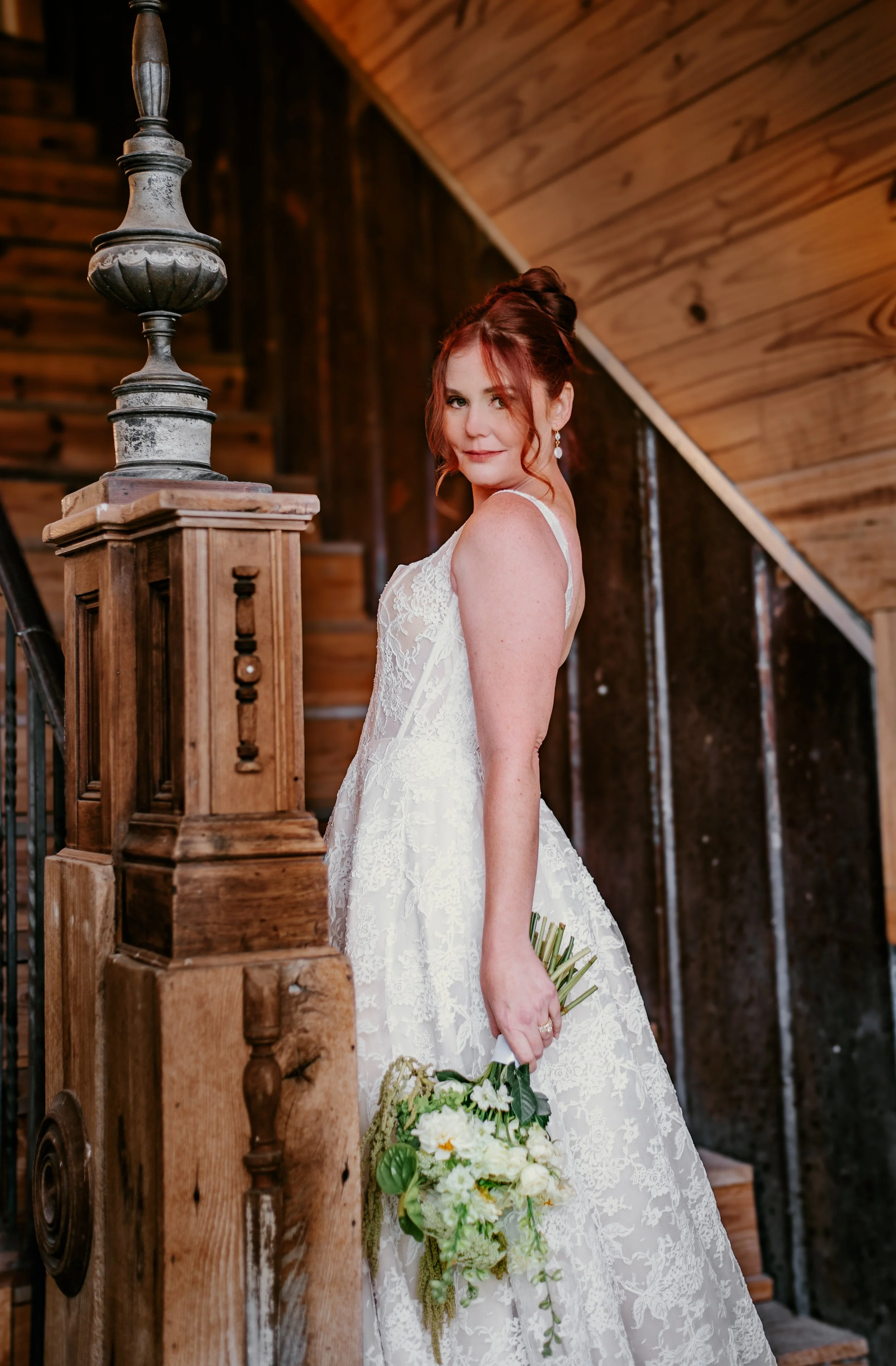 A woman in a white lace wedding dress holding a bouquet of white flowers, standing on a wooden staircase with a wooden-paneled wall behind her.