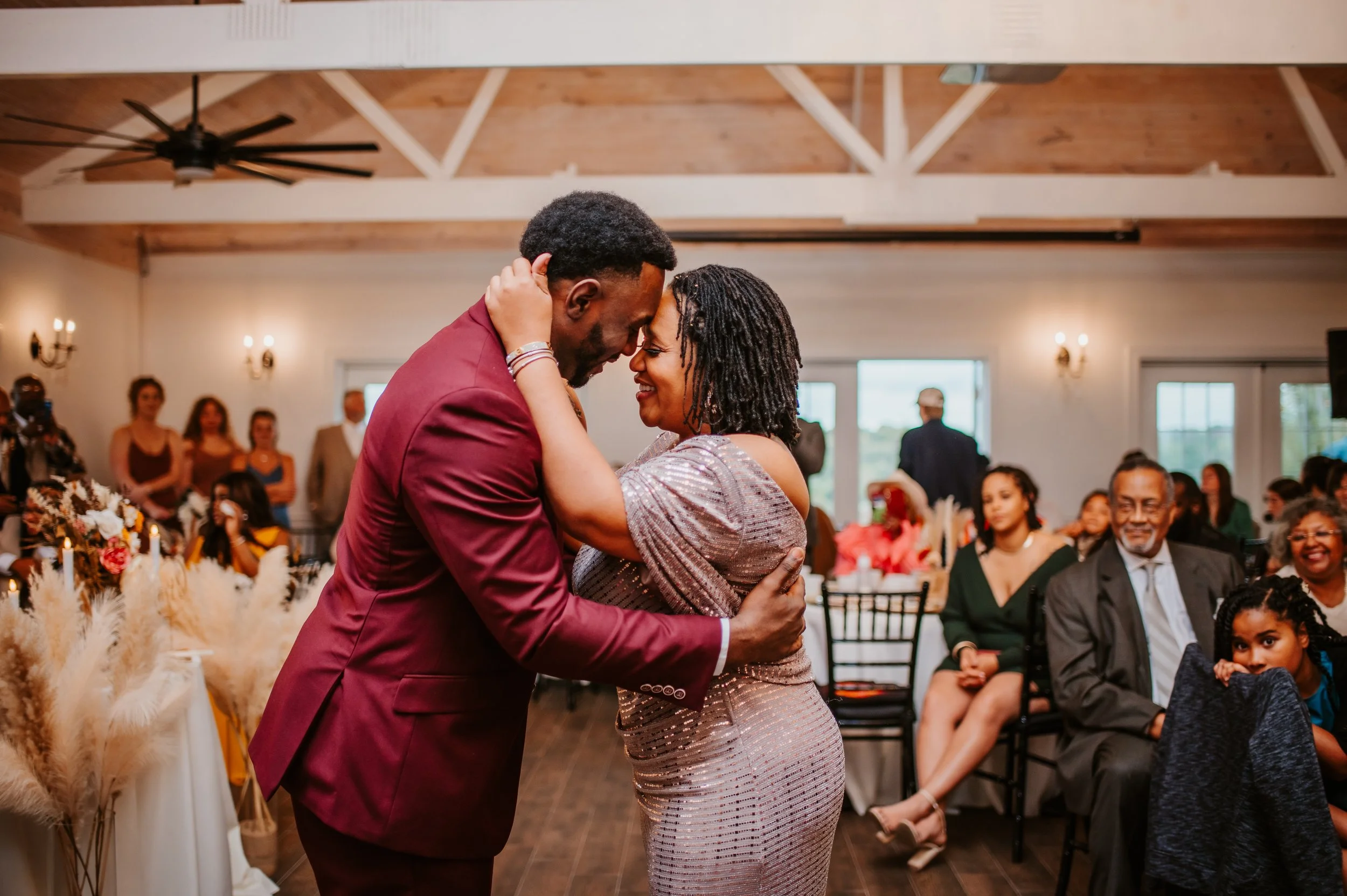 A couple is dancing closely at a celebration, smiling, surrounded by seated and standing guests in a warmly lit room with wooden ceiling beams and large windows.