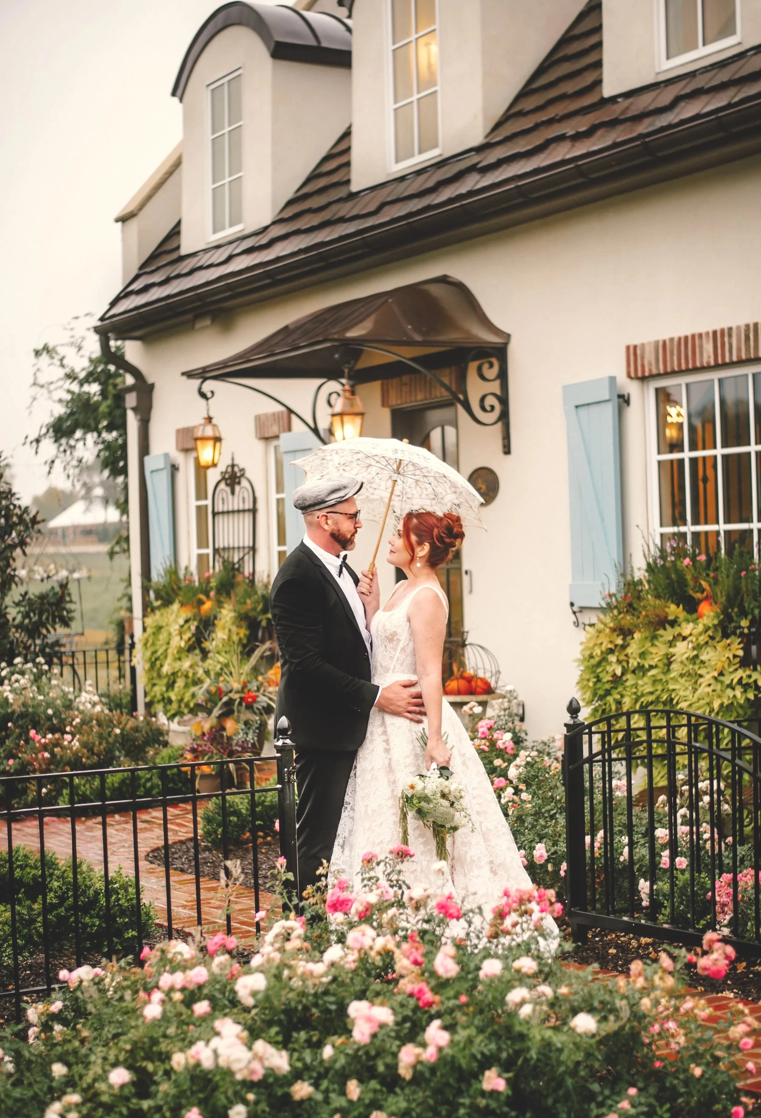 A bride and groom standing close together outside a white house with blue shutters, surrounded by pink and white flowers, sharing an intimate moment under a small umbrella on a rainy day.