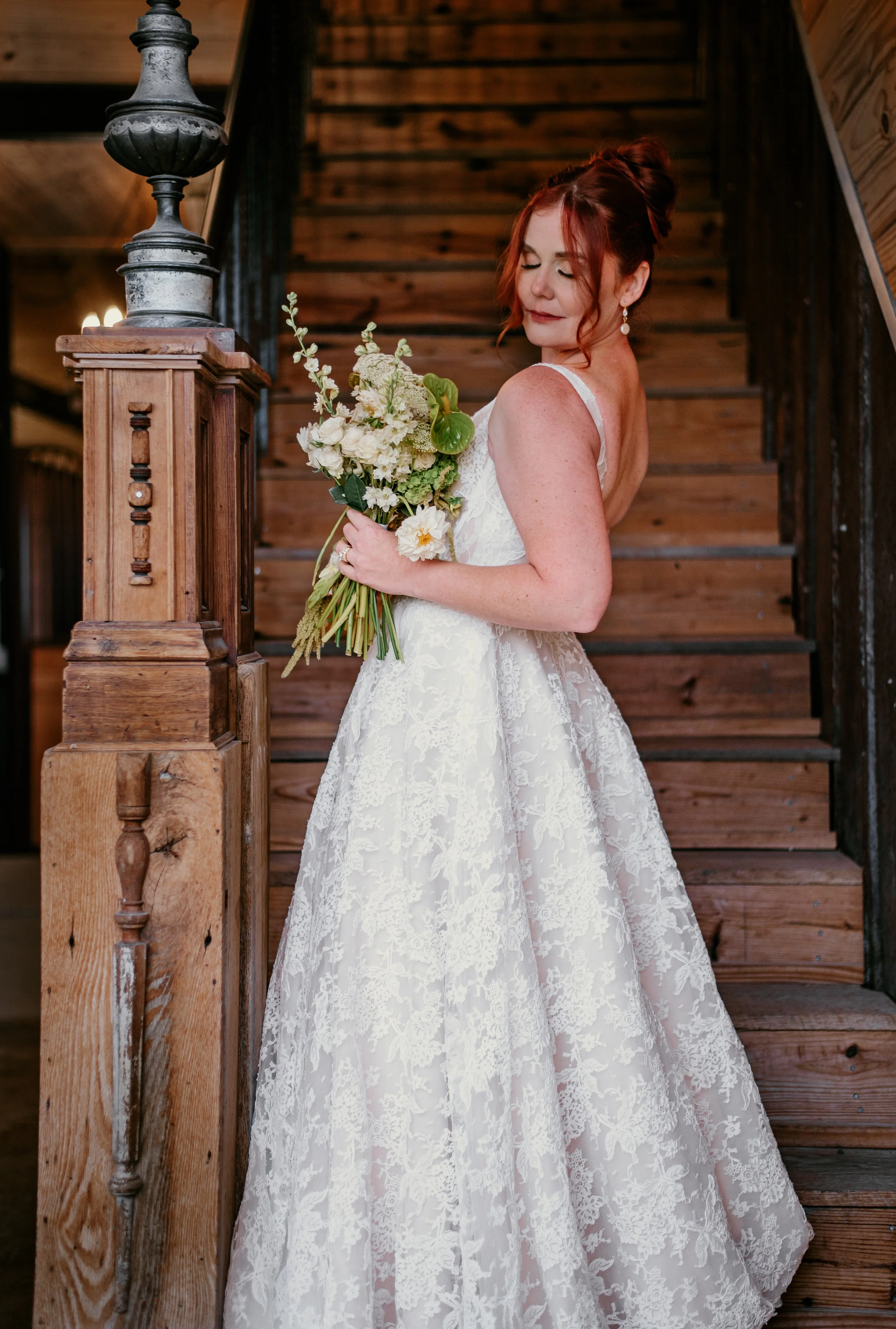 A bride with reddish-brown hair styled in an updo, wearing a white lace wedding gown with spaghetti straps, holding a bouquet of white flowers and green foliage, standing on a wooden staircase inside a rustic venue.