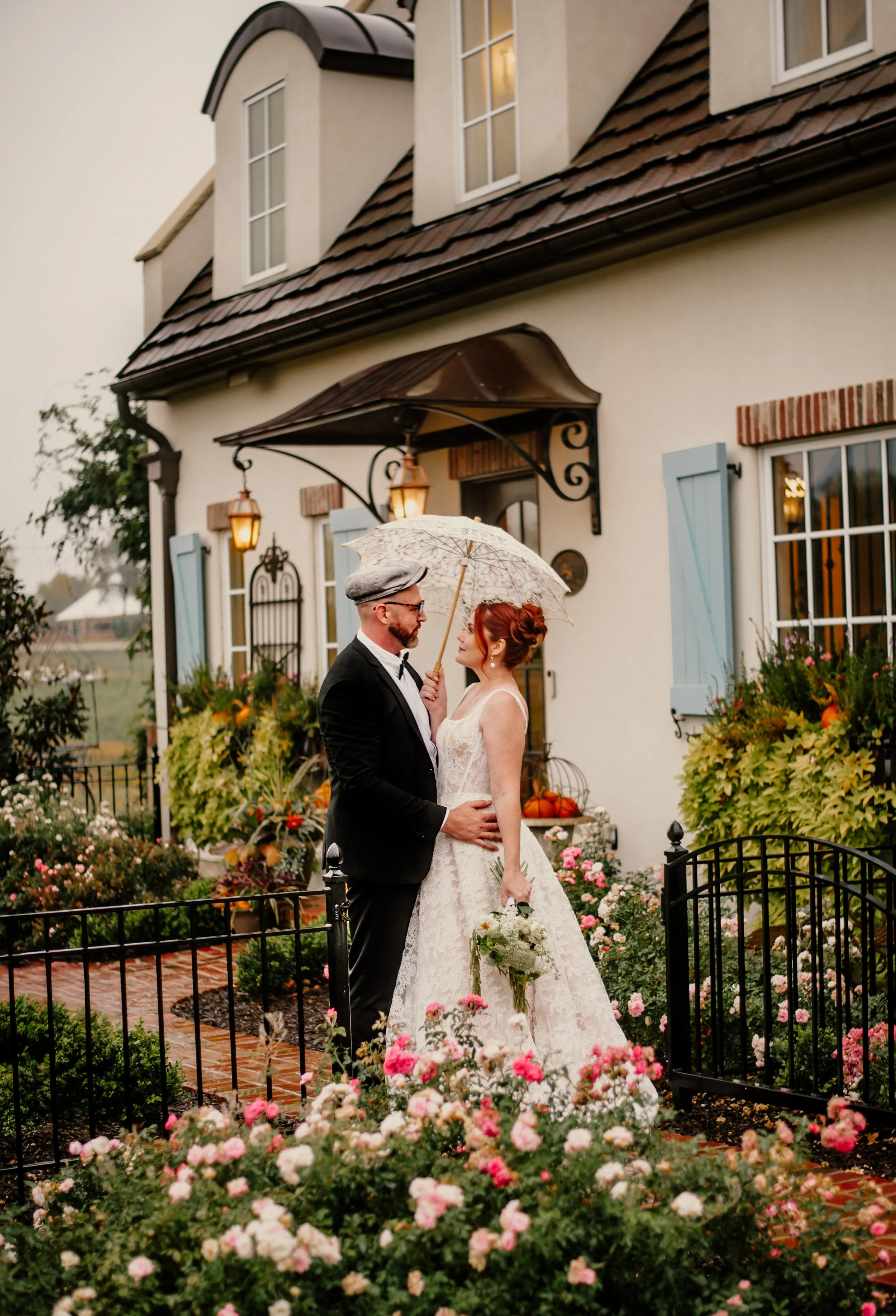 A bride and groom stand close together in a garden in front of a white house with blue shutters and a brown tiled roof, holding a clear umbrella. The bride wears a white lace wedding dress with a bouquet, and the groom wears a black tuxedo with a bow