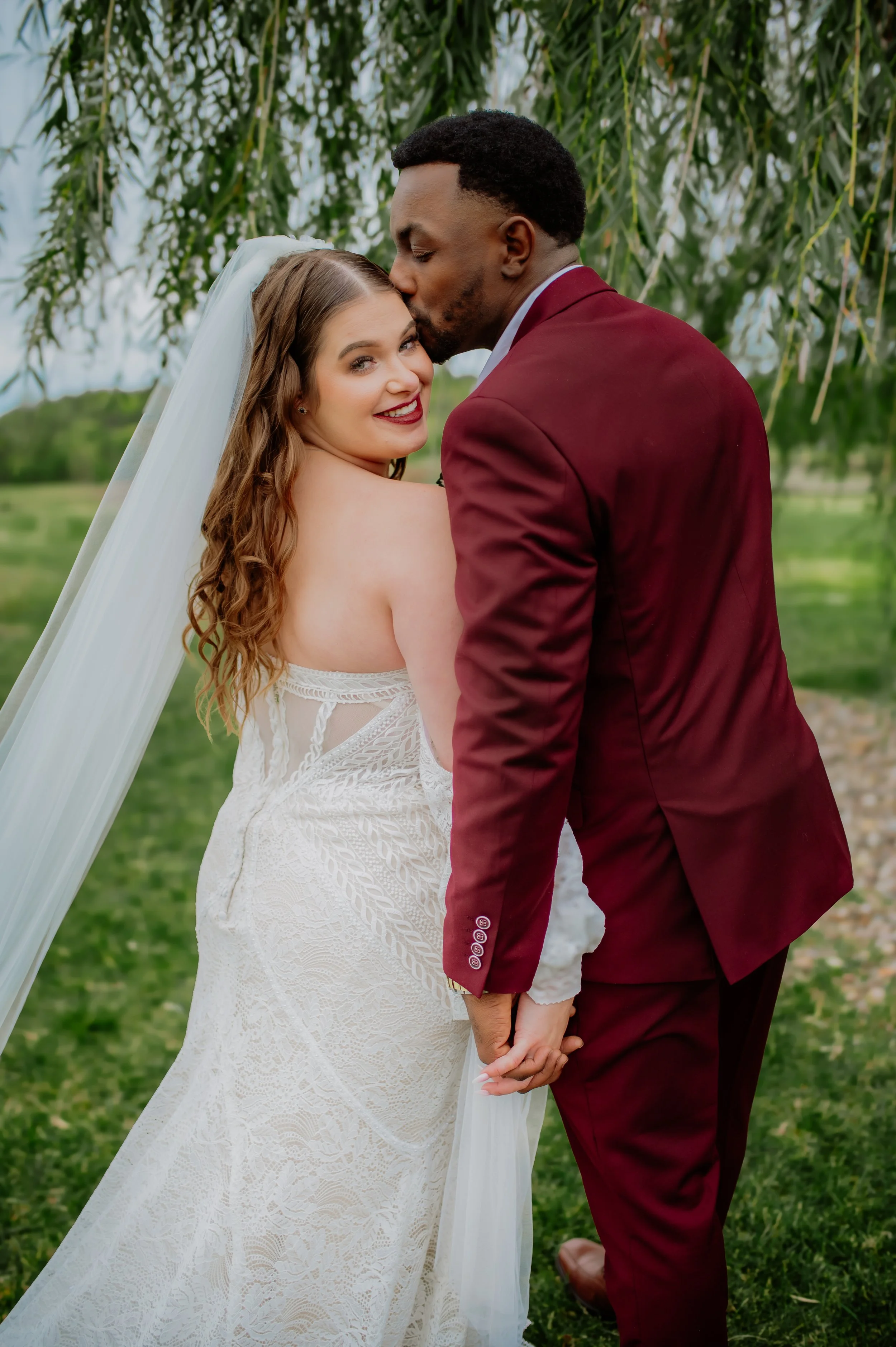 A bride with long, curly hair wearing a white lace wedding dress and veil, holding hands with a groom in a burgundy suit, kissing her on the forehead outdoors in front of green trees.