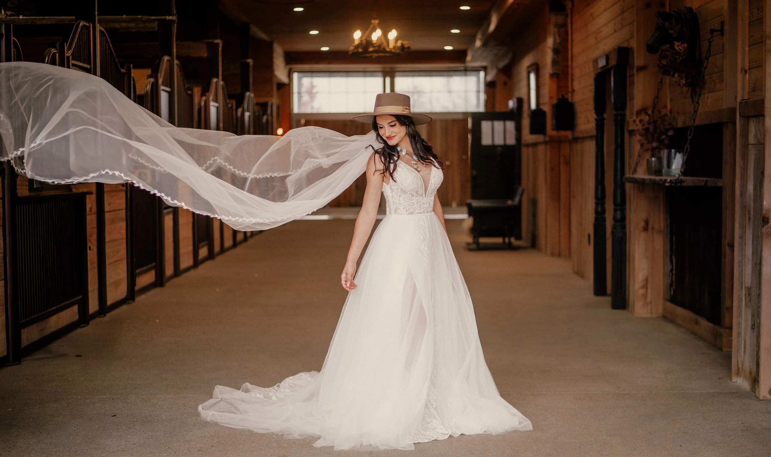 A woman in a white wedding dress and wide-brimmed hat with a veil inside a wooden building with stalls.