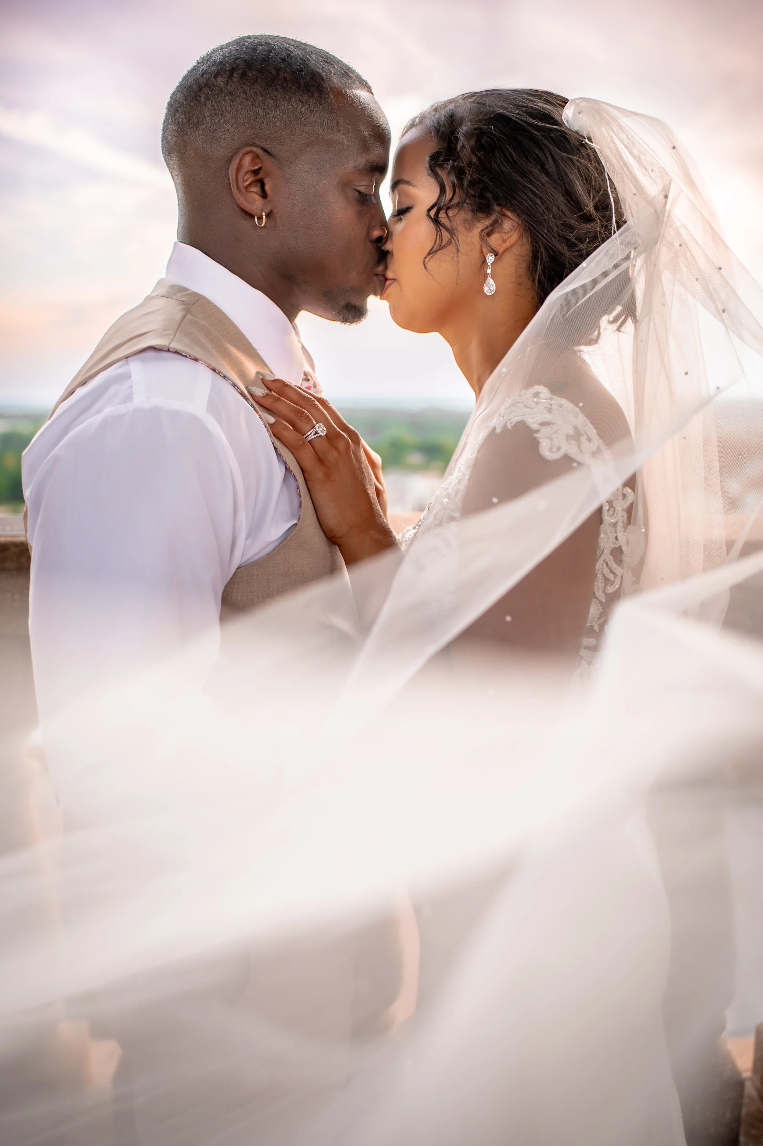 A couple dressed in wedding attire sharing a kiss outdoors, with a sunset sky and landscape in the background.