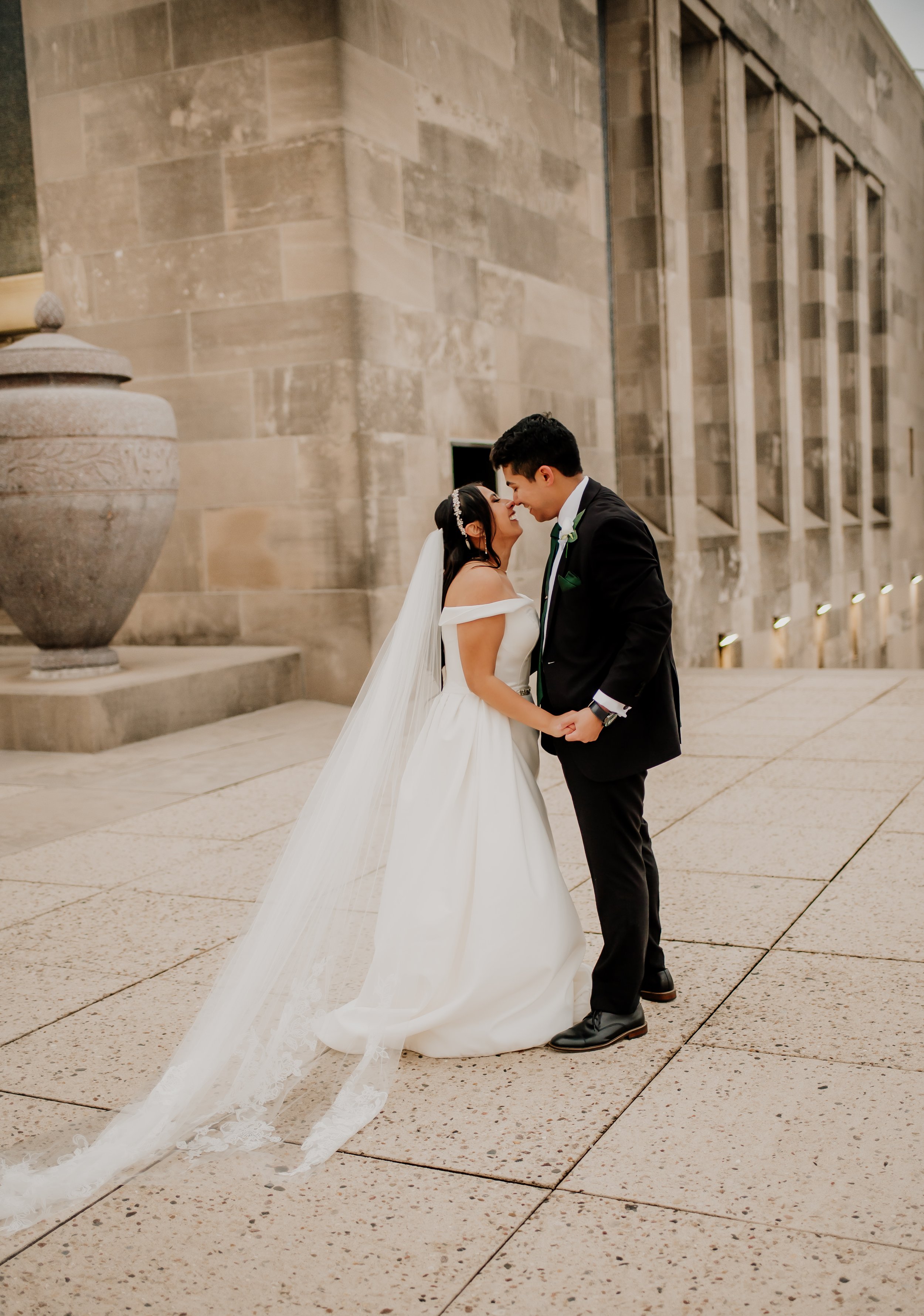 A bride and groom sharing a moment of intimacy, holding hands and smiling at each other, outside a building with modern architecture and large stone urns.