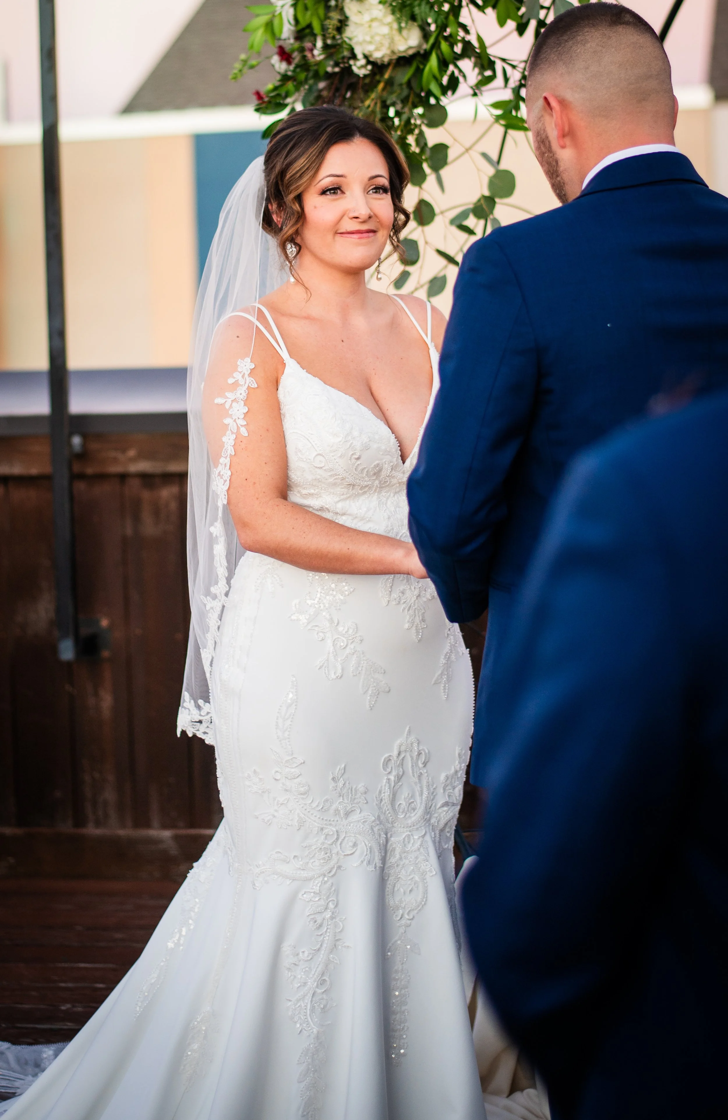 Bride with brown hair in wedding dress with lace details, veil, and earrings, facing groom in blue suit with shaved head, during wedding ceremony outdoors under floral arch.