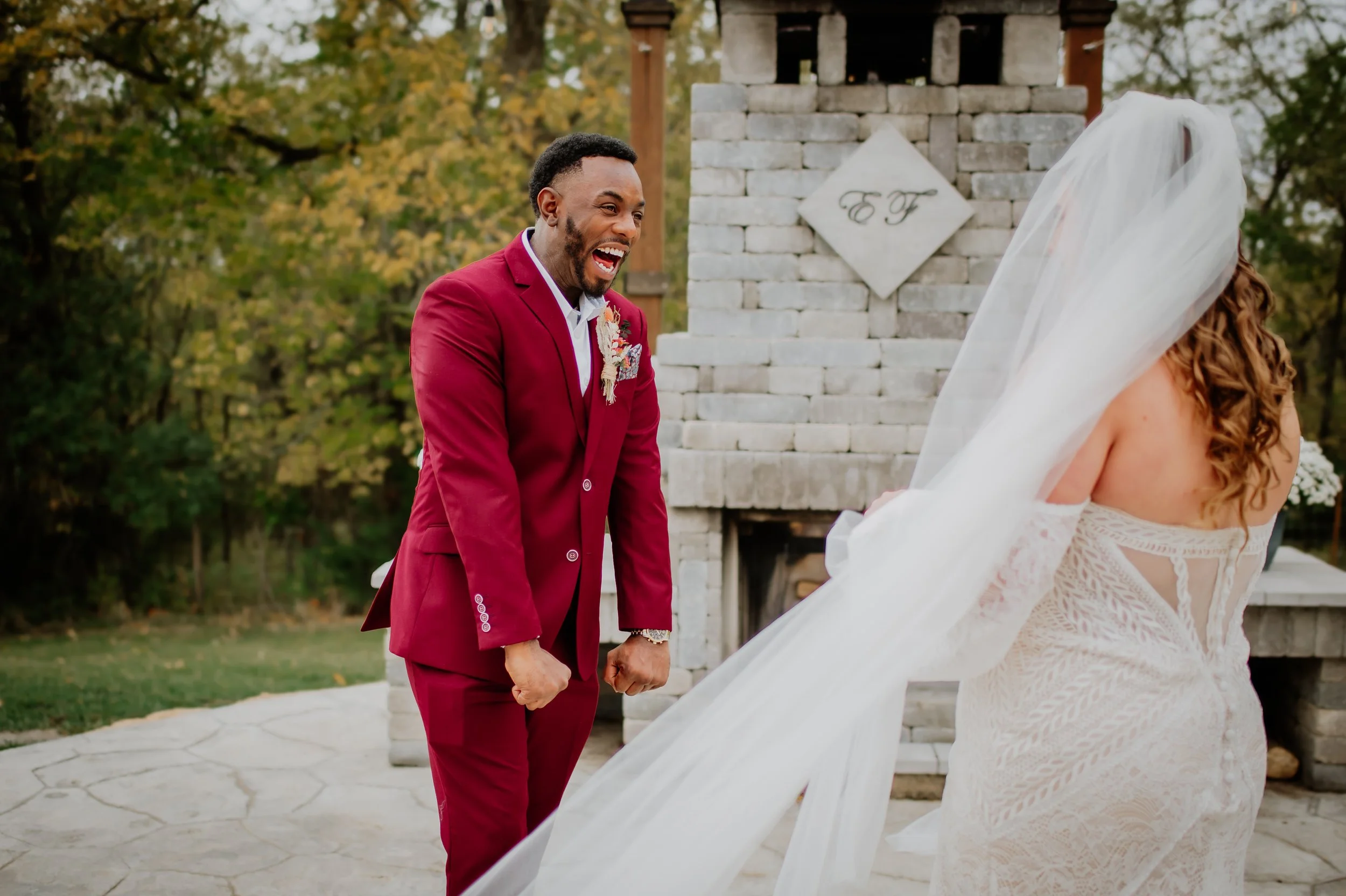 A man in a red suit smiling and holding hands with a bride in a white lace wedding dress and veil, during a wedding ceremony outdoors with trees and a white brick structure in the background.