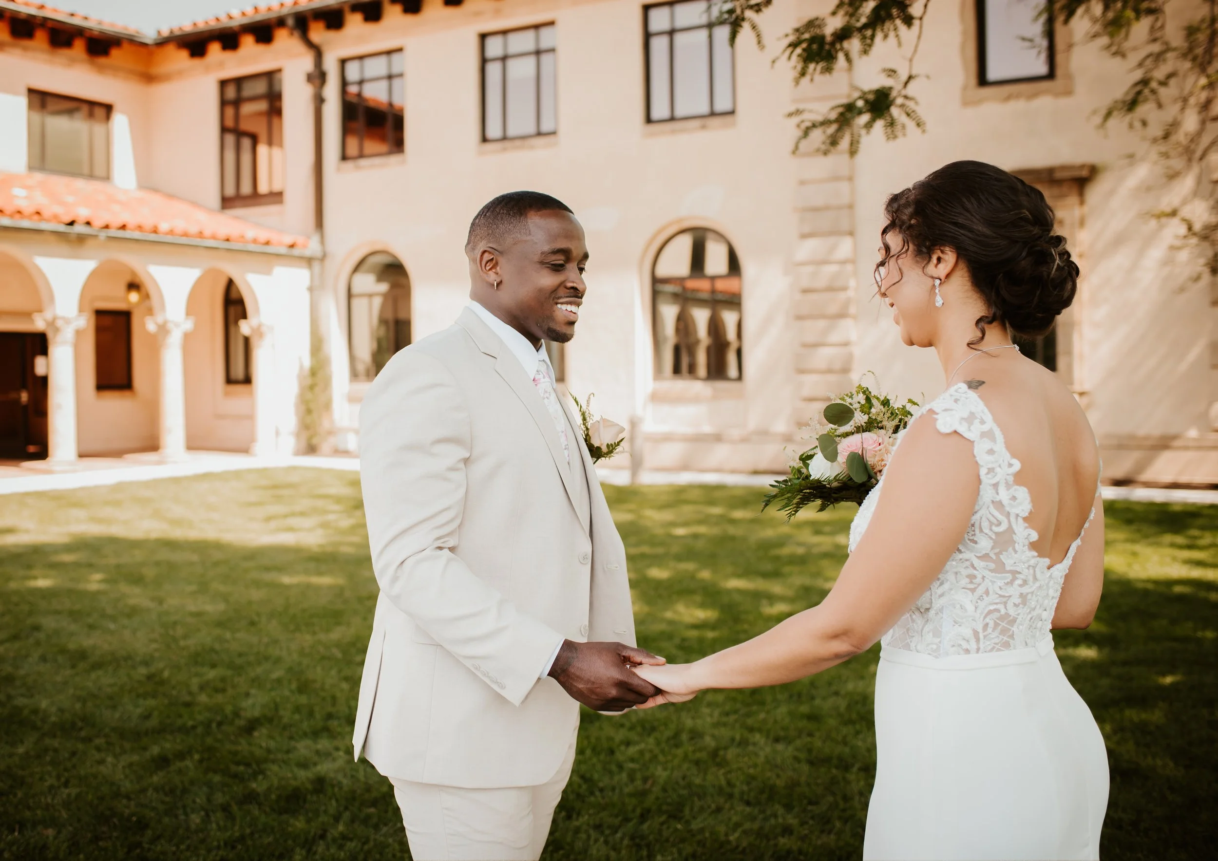 A bride and groom standing outdoors in front of a large building, holding hands and gazing at each other during their wedding ceremony.