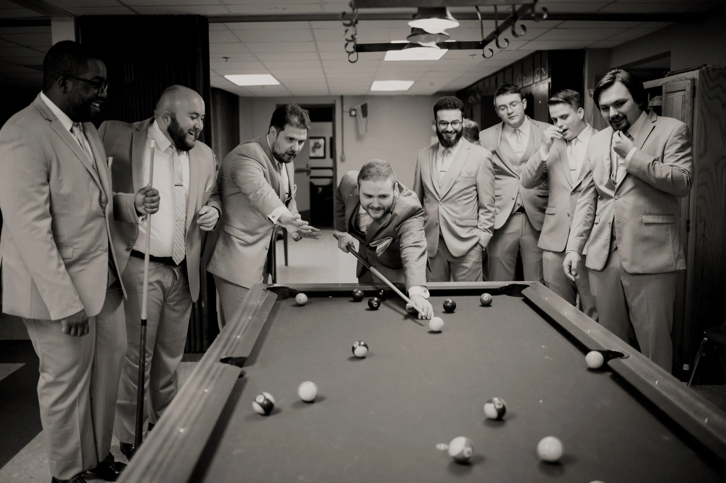 A group of men in suits standing around a pool table, watching a game. One man is leaning over to take a shot, while others observe with smiles and interest.