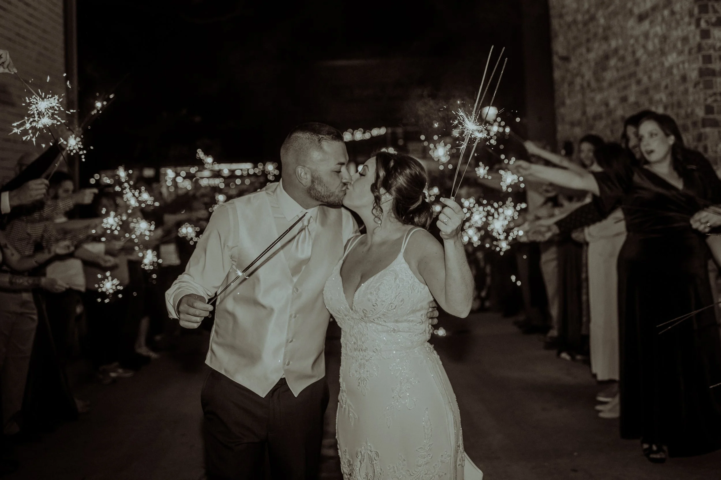 A newlywed couple shares a kiss at night during a celebration with sparklers, surrounded by friends and family.