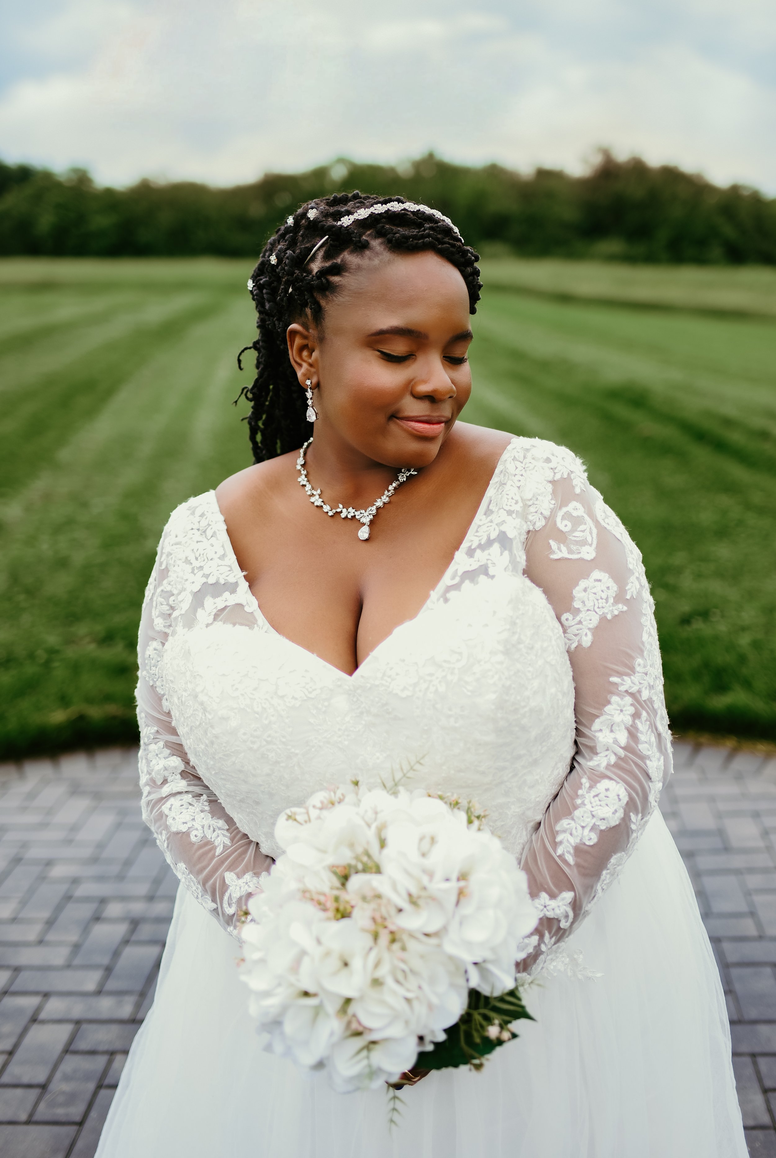 A bride in a white lace wedding dress holding a bouquet of white flowers, standing outdoors on a paved pathway with a grassy field and trees in the background, with her eyes closed and a gentle smile.