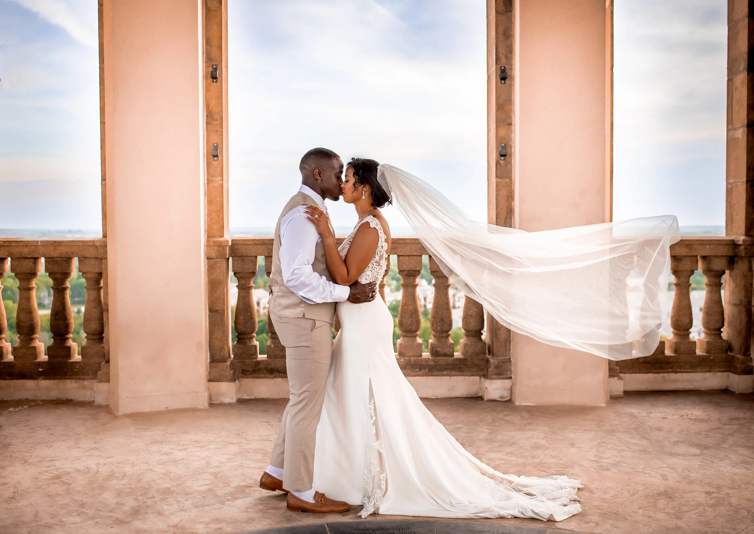A bride and groom share a kiss on a balcony with a scenic view of the sky and landscape behind them.