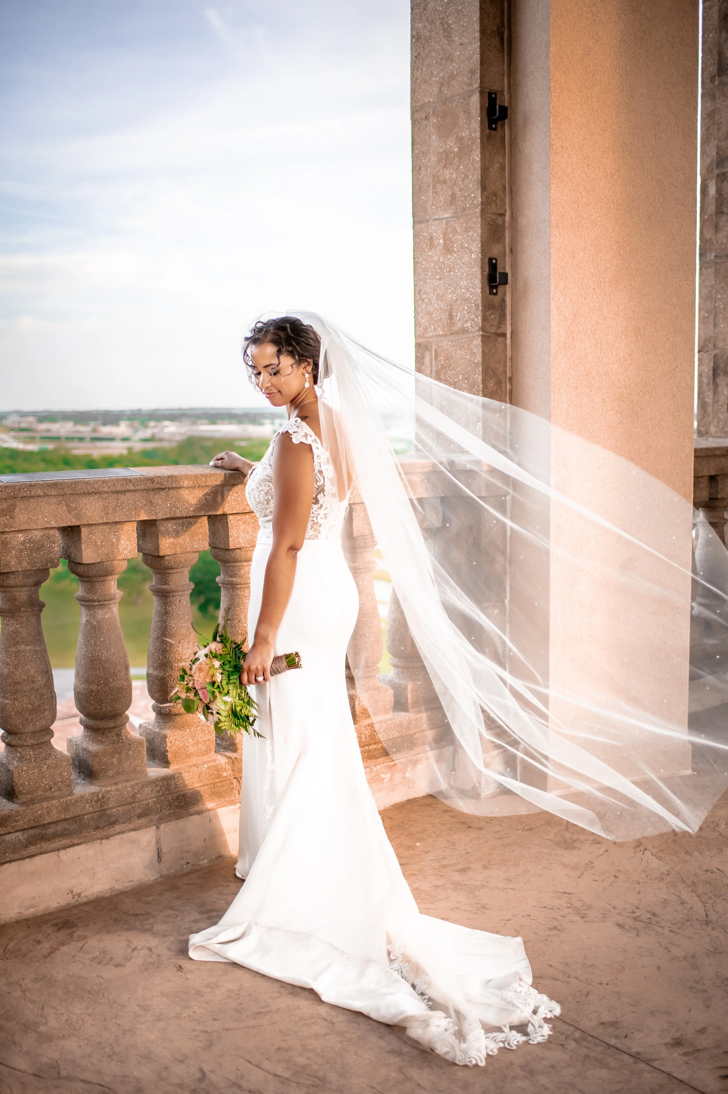 A bride in a white wedding gown with lace details on the shoulders, holding a bouquet of flowers, standing on a balcony with stone balustrades, with a cityscape in the background and her veil flowing behind her.