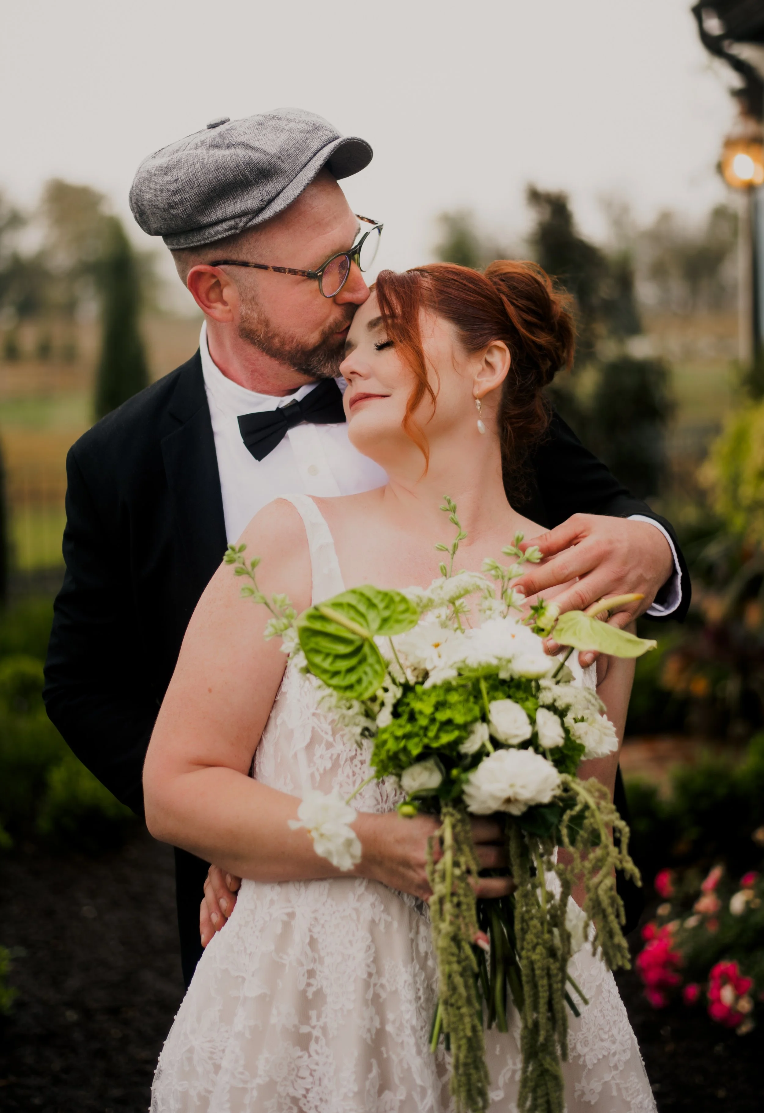 A newlywed couple embracing outdoors, the groom kissing the bride's forehead as she smiles with closed eyes, holding a large bouquet of white and green flowers.