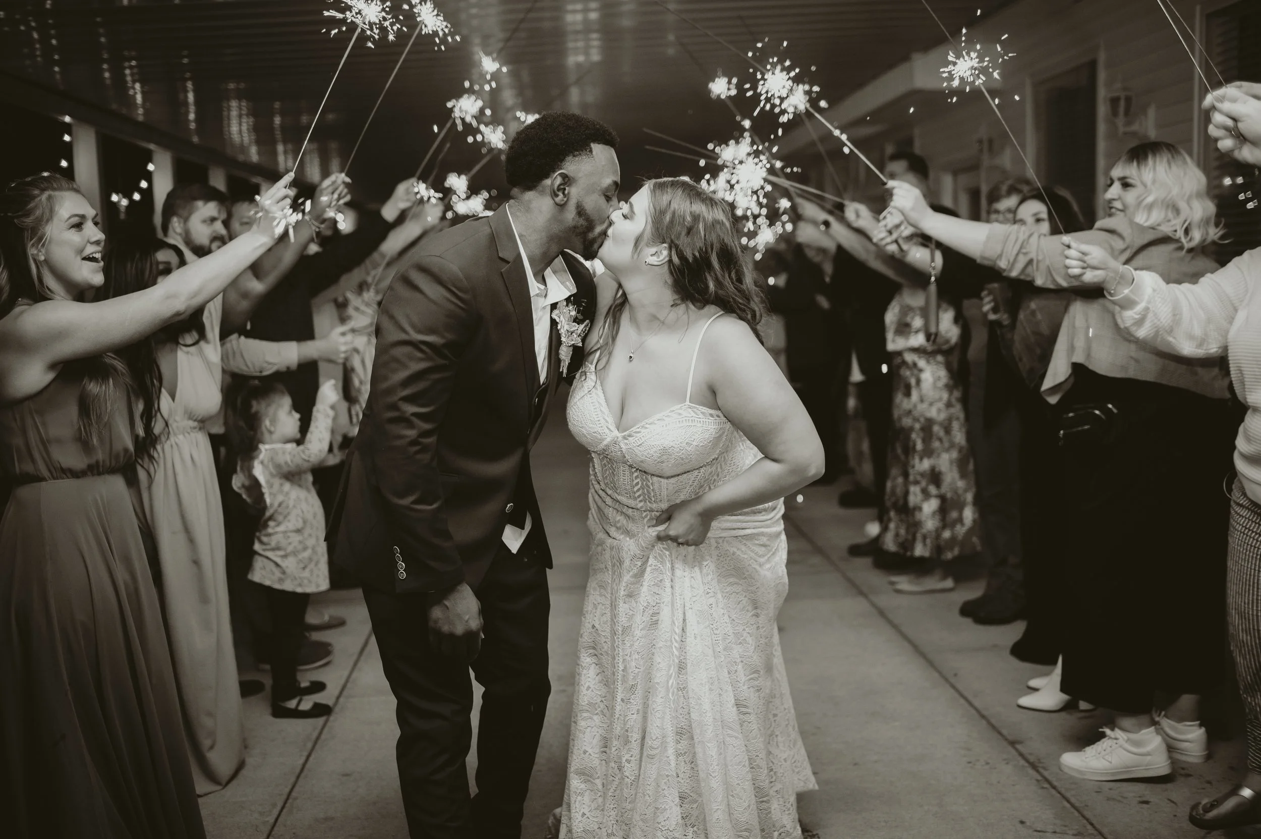 A newlywed couple kissing in the center of a dance floor surrounded by guests holding sparklers at night.