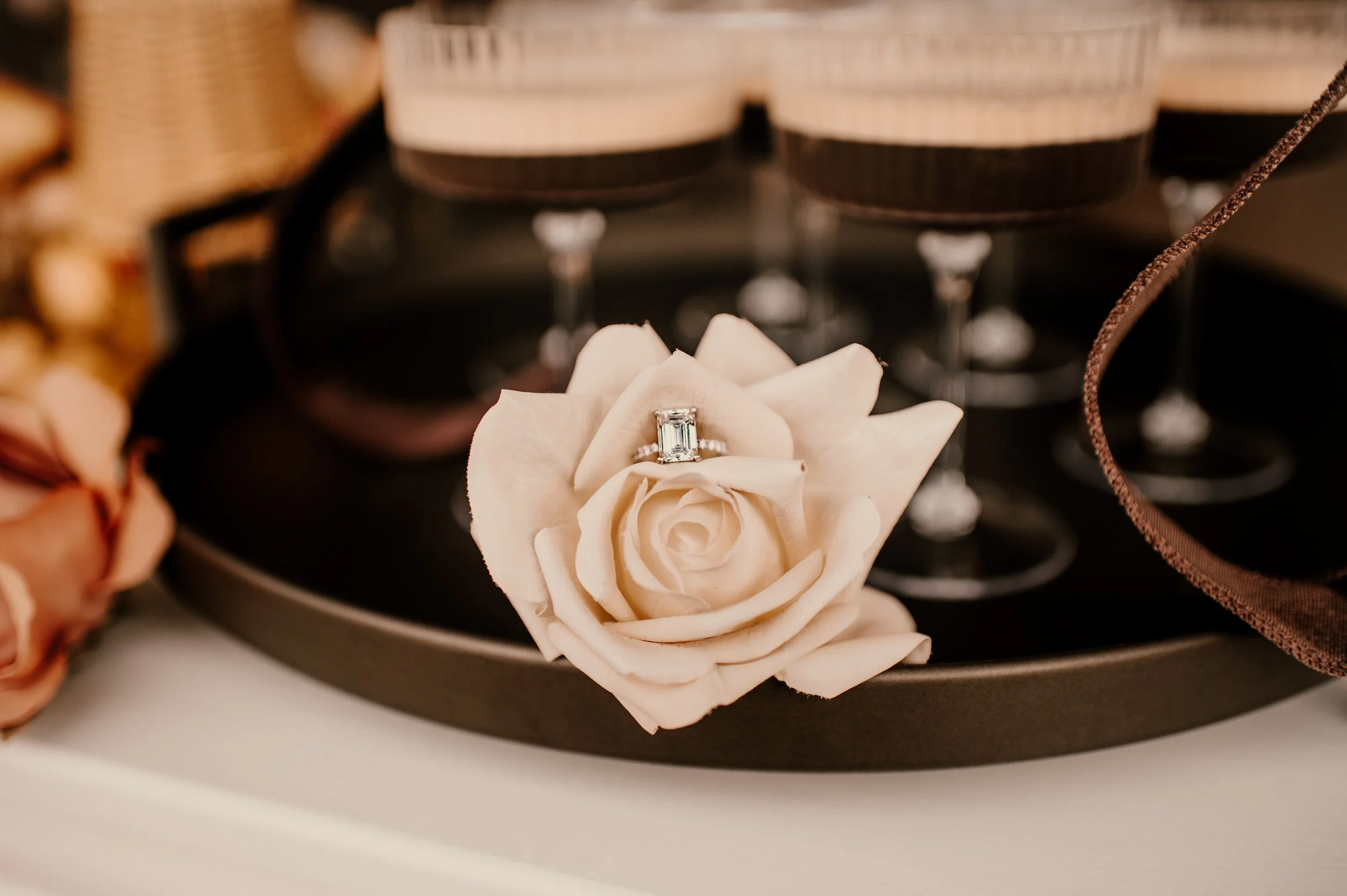 A diamond engagement ring on a light peach-colored rose, placed on a black tray with other roses and jewelry items in the background.
