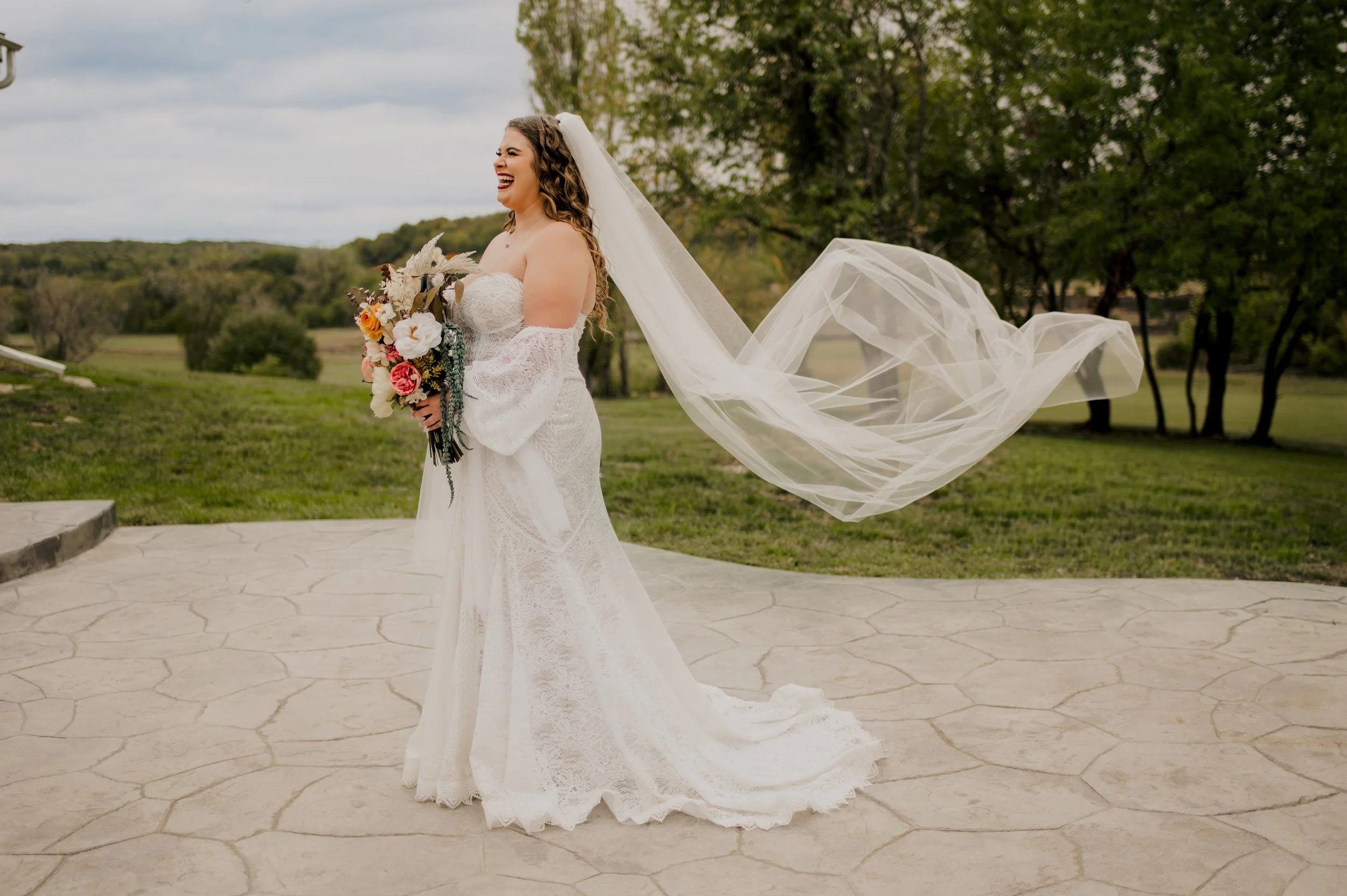 A bride in a white wedding dress holding a bouquet of flowers, standing outdoors on a paved surface with green grass and trees in the background, smiling and laughing.