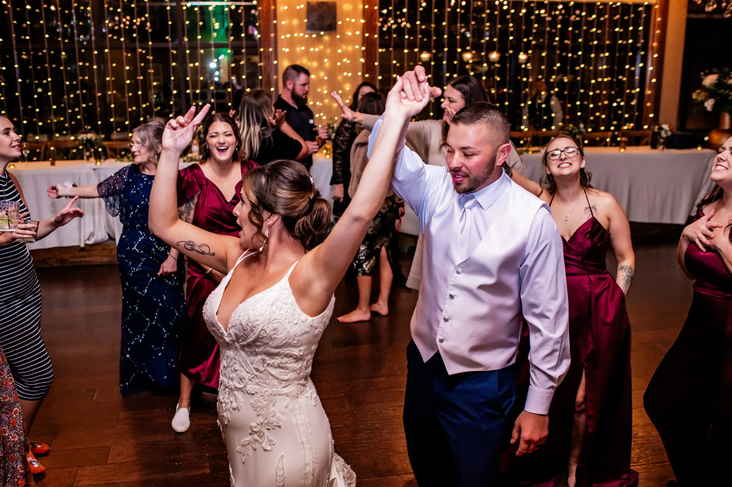 People dancing and celebrating at a wedding reception with warm string lights in the background.