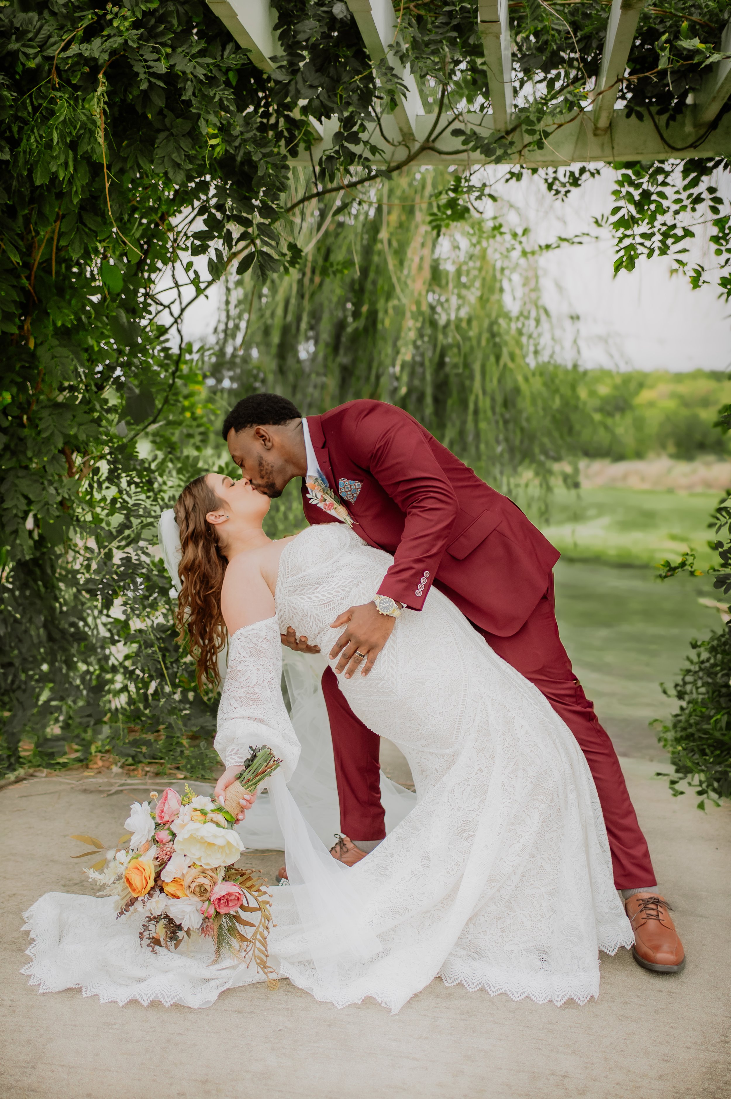 A bride and groom sharing a kiss outdoors during their wedding, with the groom dipping the bride, surrounded by greenery, with a bouquet of flowers on the ground.