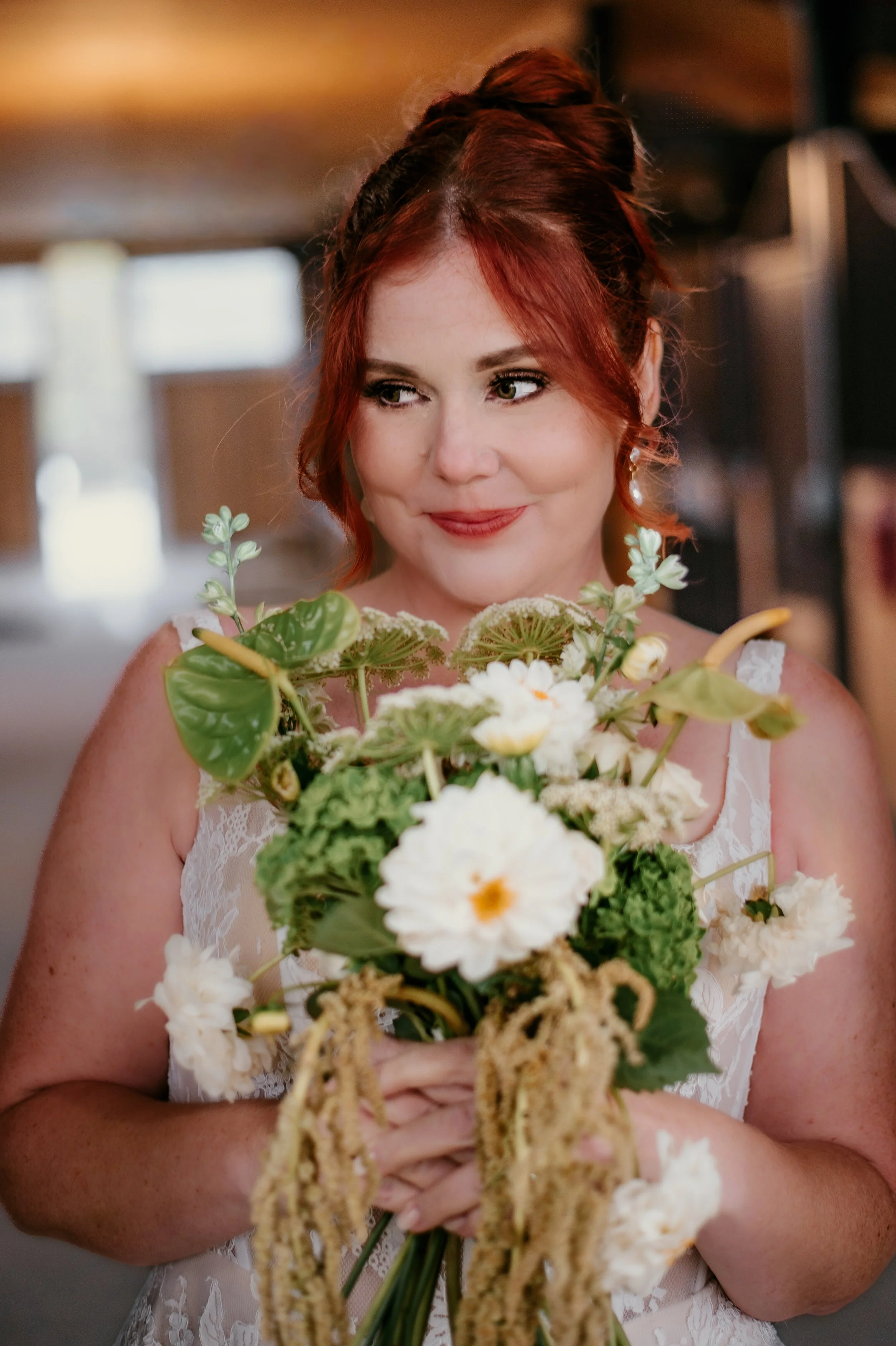A woman with red hair in an updo holding a bouquet of white flowers and green foliage, wearing a white lace dress, standing indoors with a blurred background.